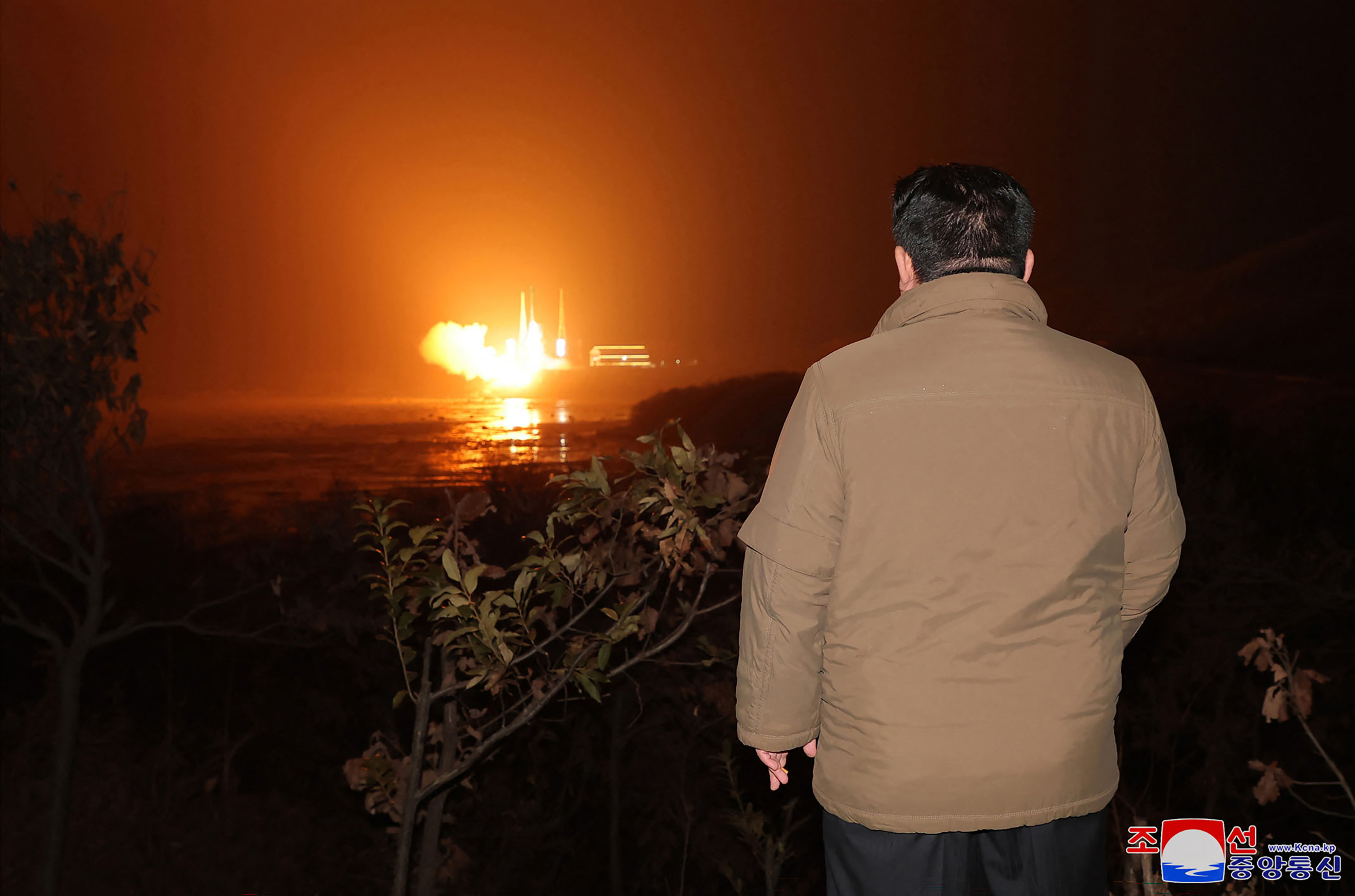 Kim Jong Un observing the rocket launch. He is wearing a brown jacket and has his back to the camera. There is an orange ball of flame lighting up the night sky.