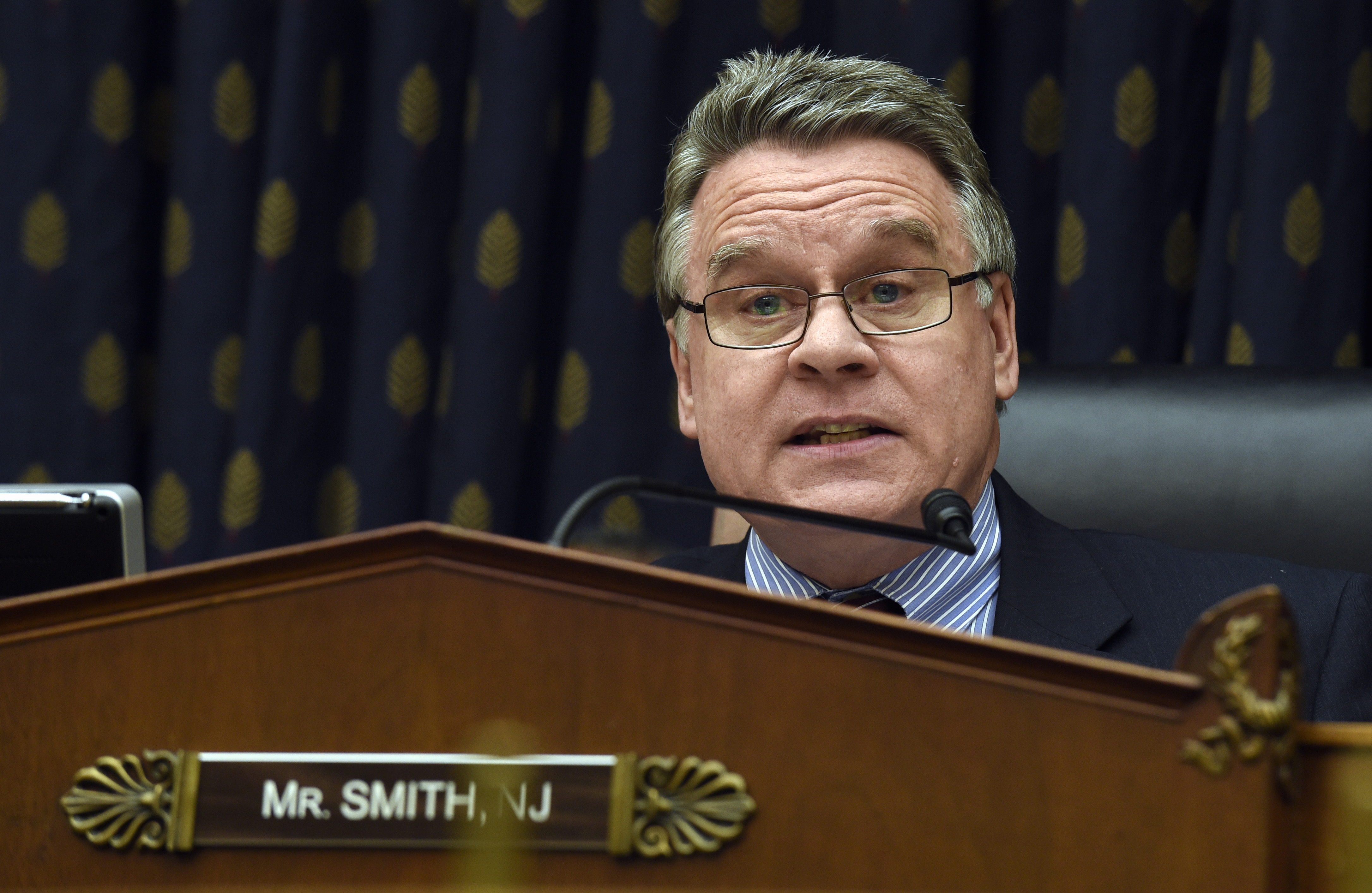 Representative Chris Smith sits behind a wooden panel with his name and the state of New Jersey indicated on a panel. He wears glasses and speaks into a microphone.