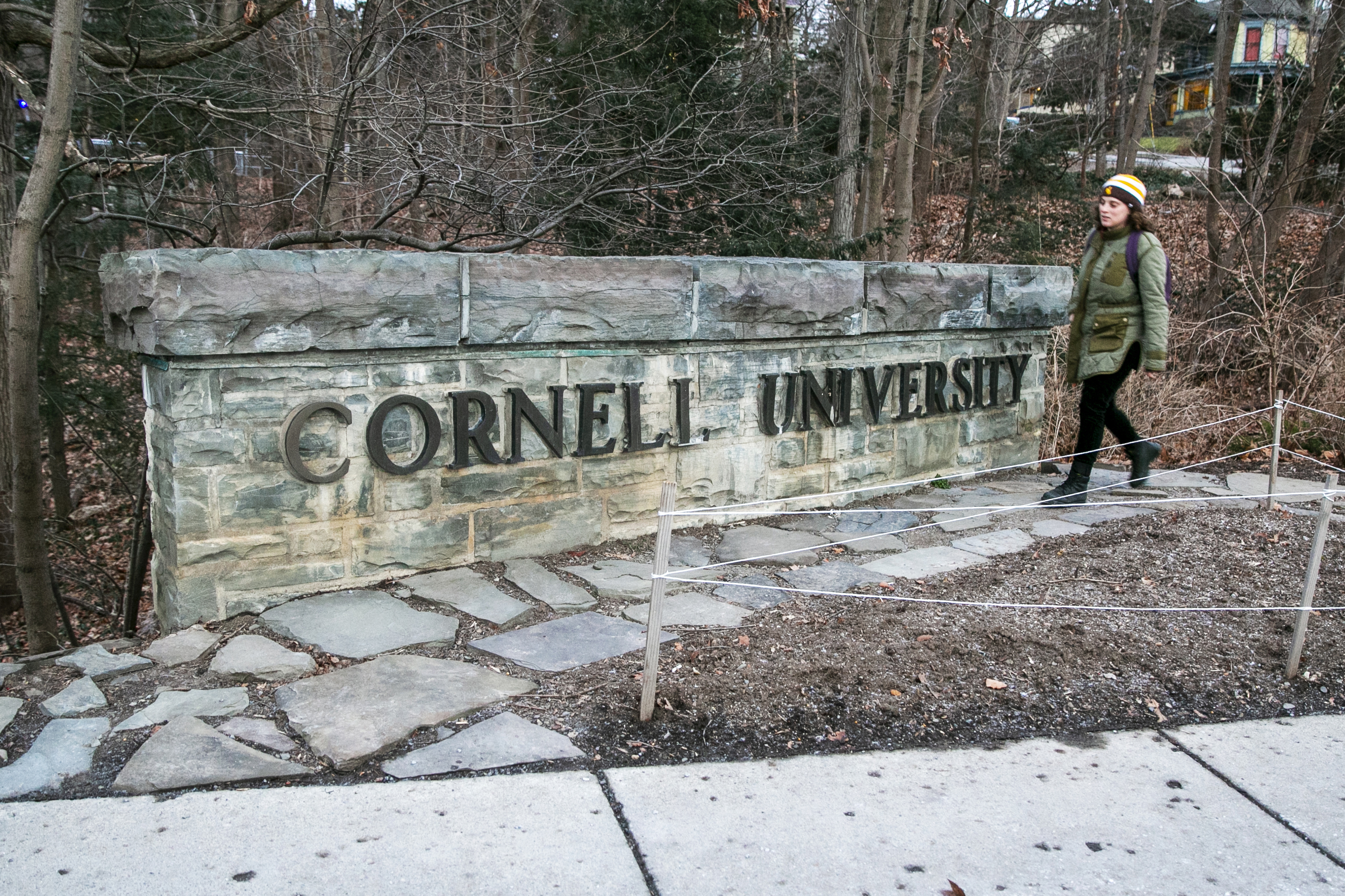 A woman walks in front of a short brick sign, with the words "Cornell University."