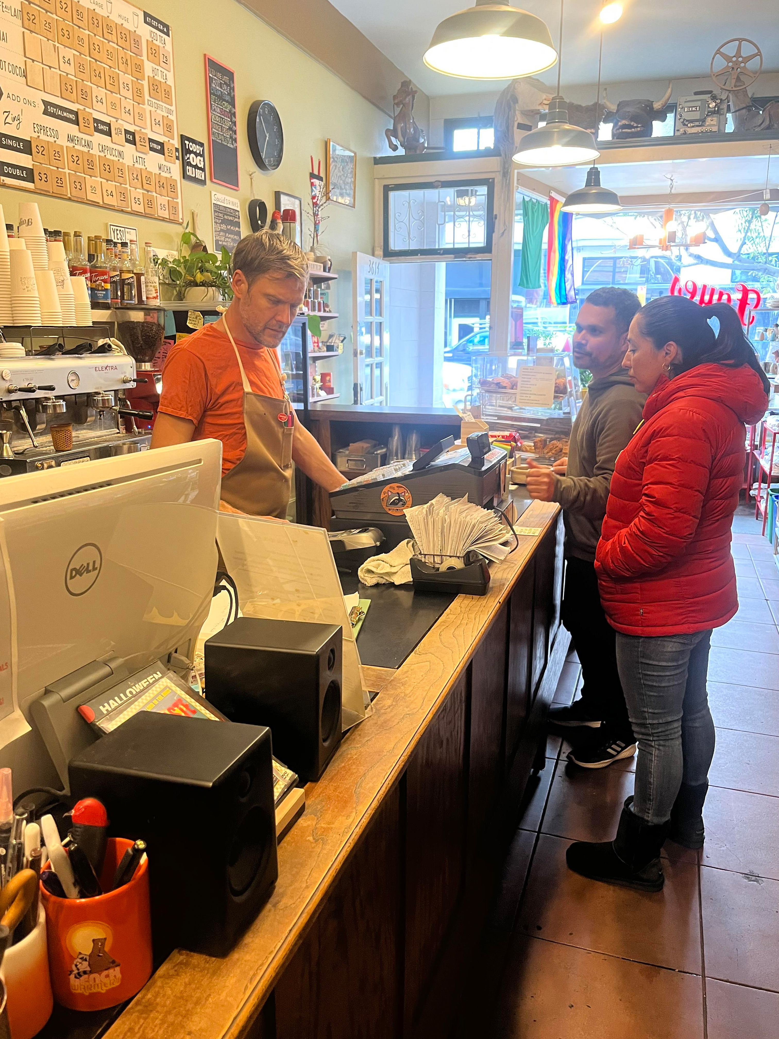 Michael McConnell, Fayes cafe co-founder, stands behind the counter of his establishment in an apron as two customers belly up to the counter.