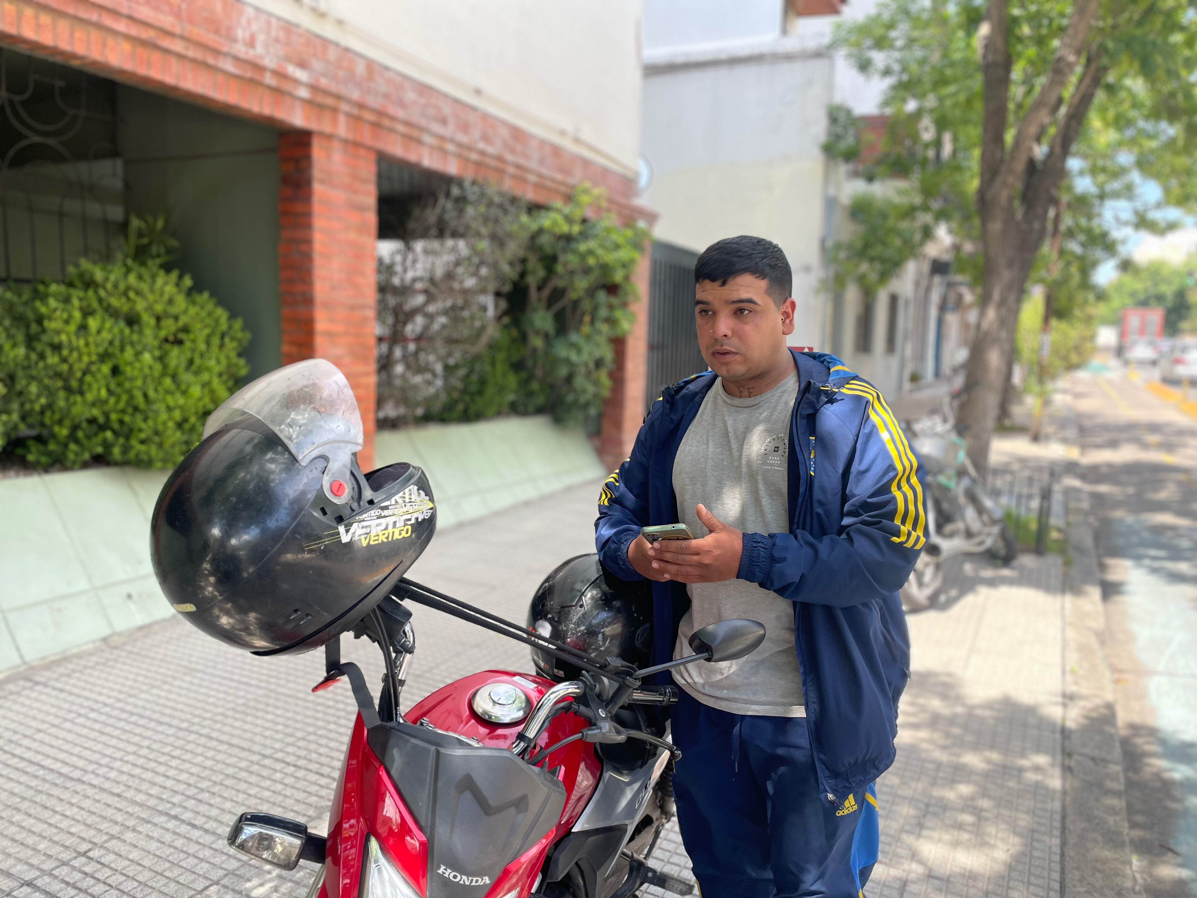 A young man, dressed in a windbreaker jacket, a T-shirt and jeans, stands beside a motorbike with a helmet propped up on the handlebars.