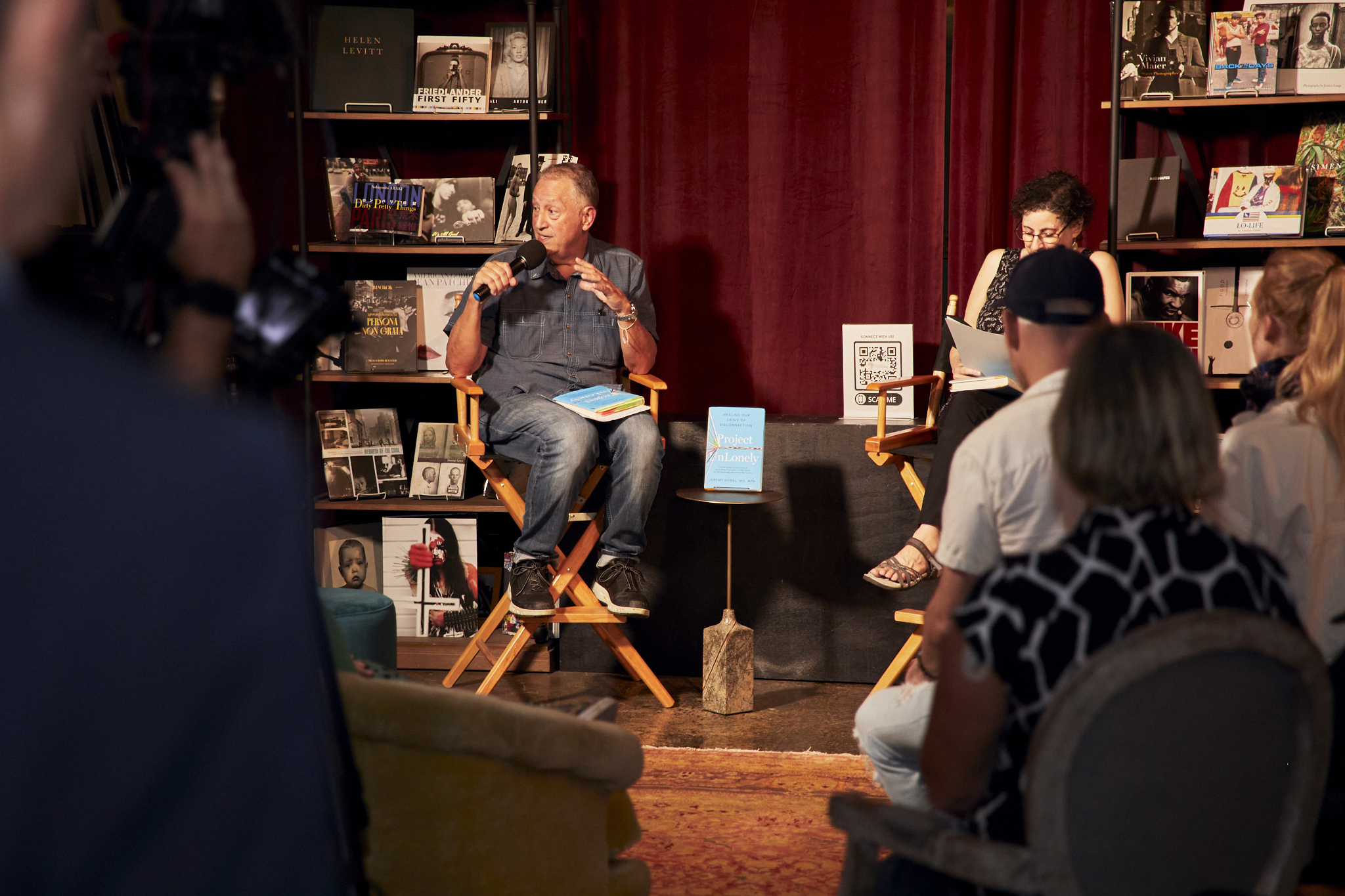 Jeremy Nobel sits in a wooden folding chair, speaking into a microphone on a small wooden stage, where he is surrounded by books. On a small table is his book, Project UnLonely.