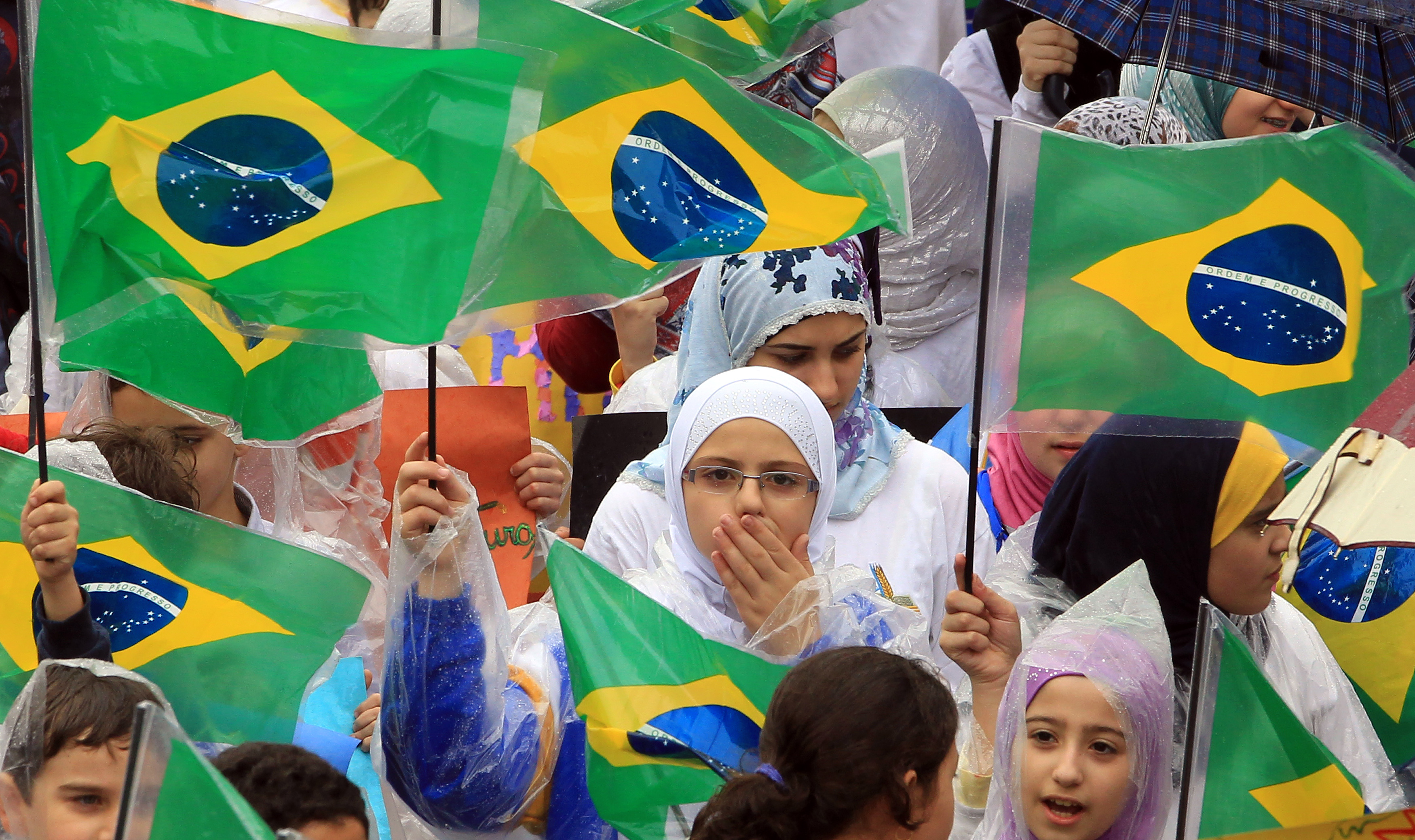 Children in hijabs wave Brazilian flags in an outdoor crowd. One girl covers her mouth with her hand.