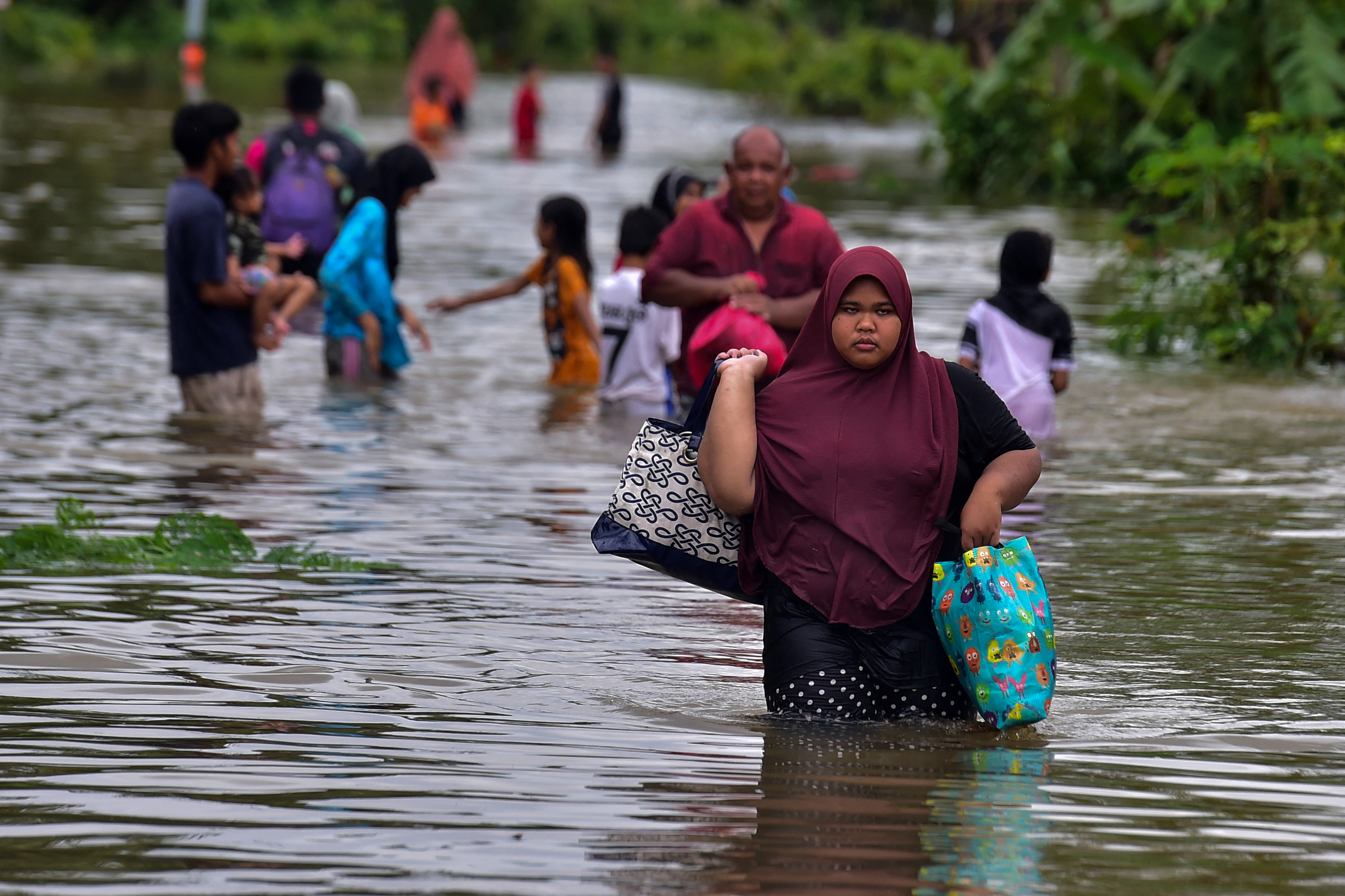 A woman leads residents along a submerged road. She is carrying some bags with her belongings. 