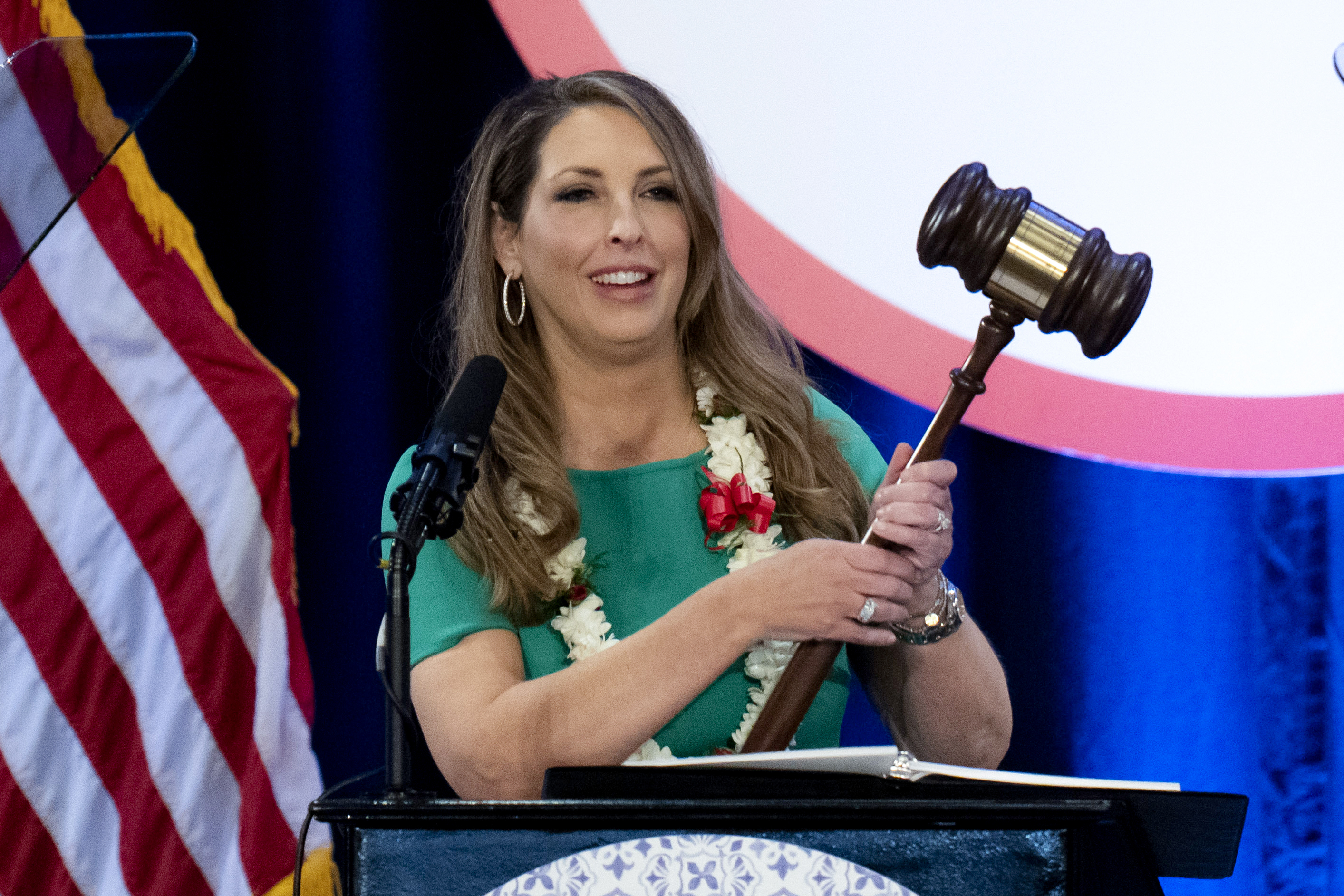 Ronna McDaniel, wearing a green shirt and a garland of flowers around her neck and carrying an oversized gavel, stands at a podium in California.