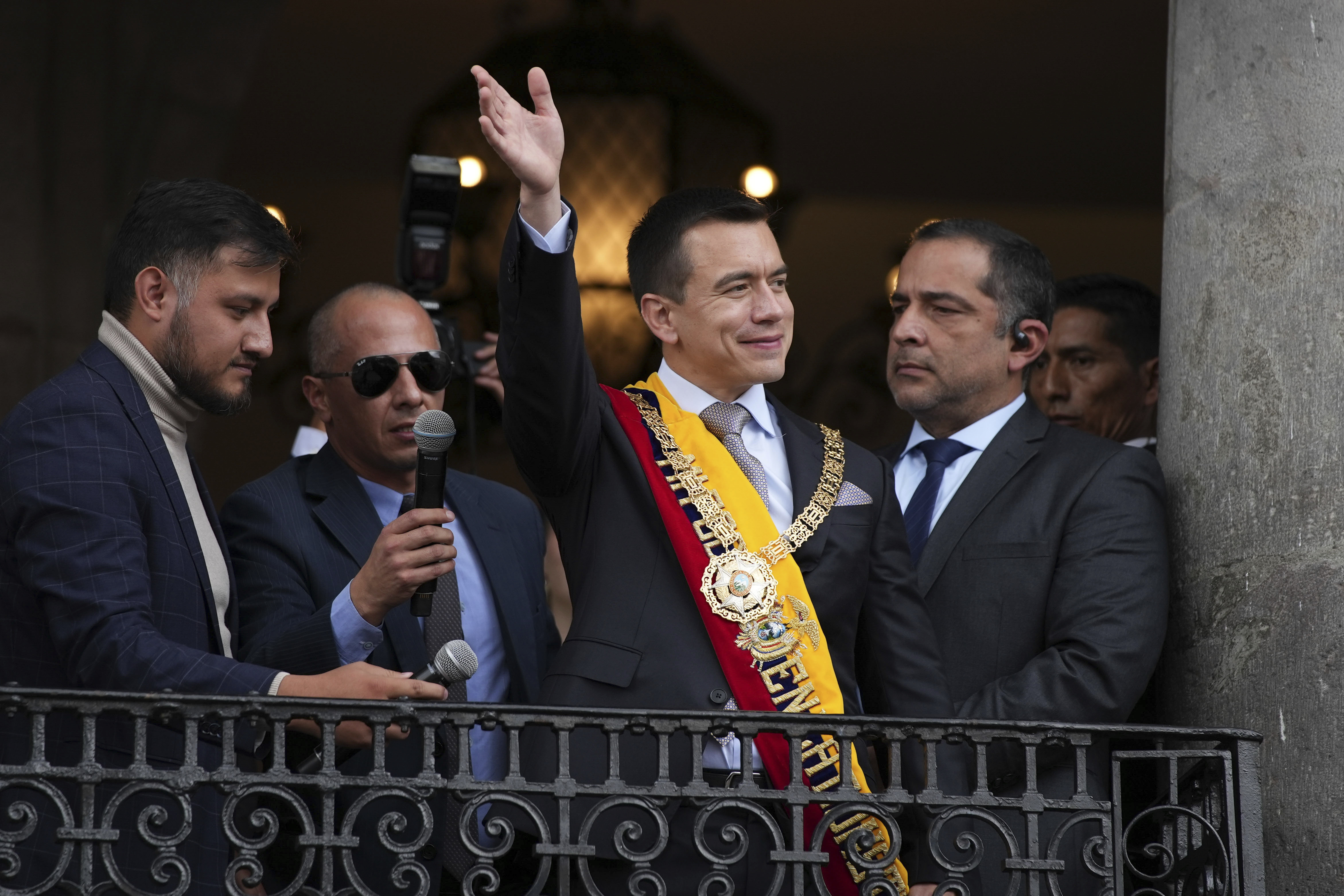 Daniel Noboa stands on a balcony with officials in dark suits, wearing a sash in the colors of Ecuador's flag and waving to the crowds below.