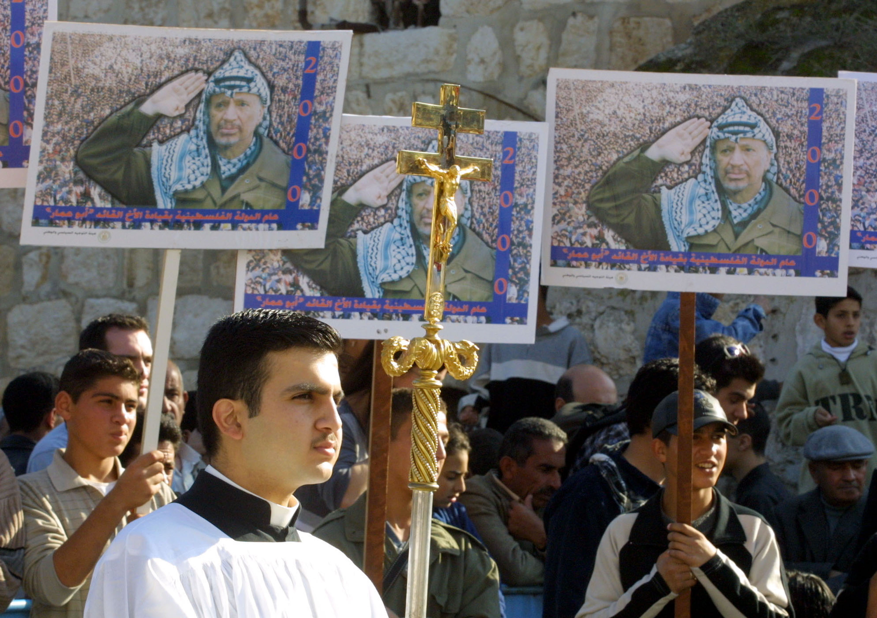 A Palestinian man holds on a golden crucifix as he waits alongside posters of Palestinian leader Yasser Arafat outside the Church of the Nativity in the West Bank town of Bethlehem December 24, 2001 during the traditional Christmas eve procession.