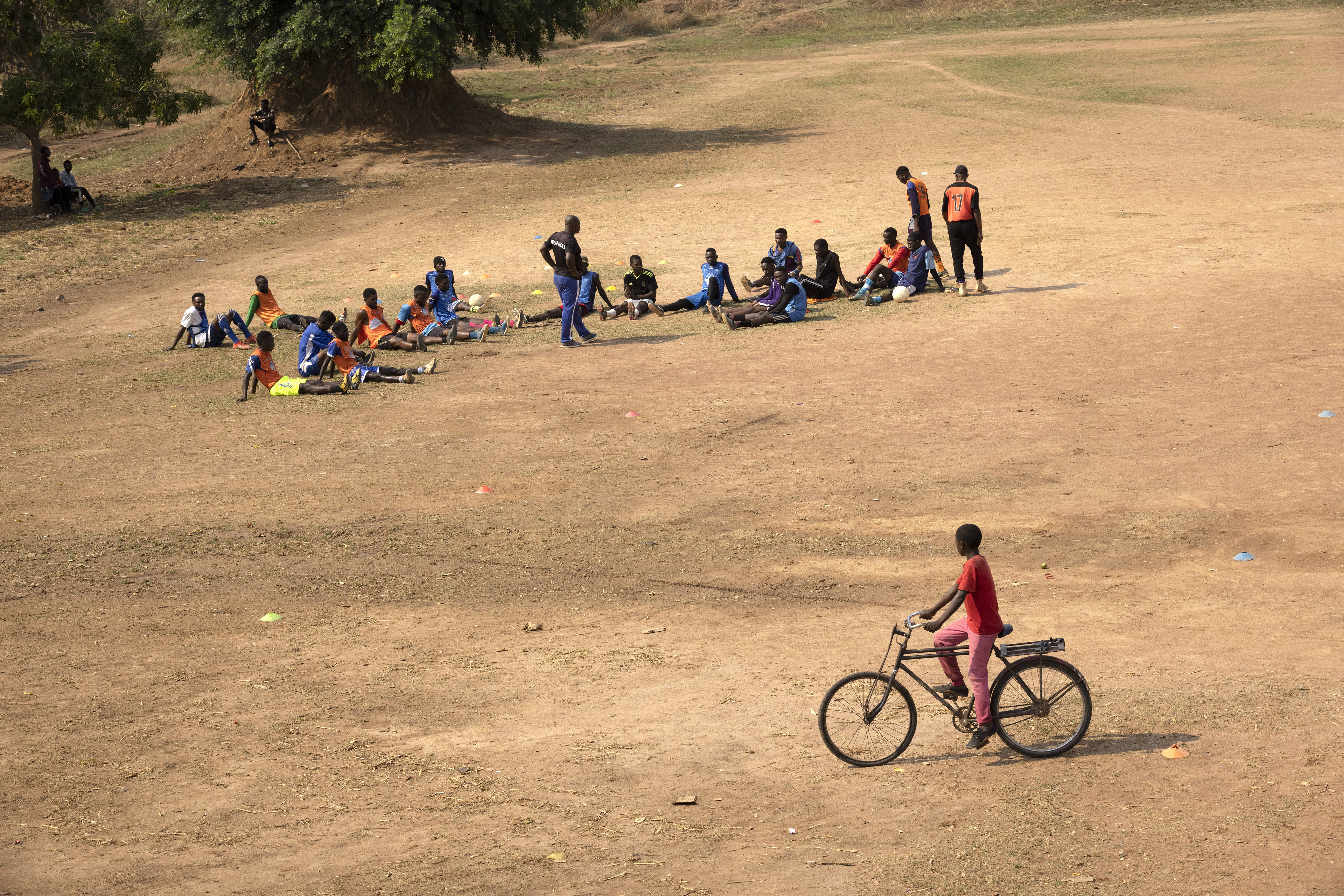 A child on a bicycle passes by as a football team gather for training on a field in a refugee camp in Zambia