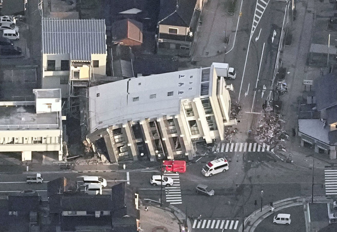 An aerial view of a multi-storey building fallen onto its side in Wajima. The building has fallen on the road. A red fire truck is nearby.