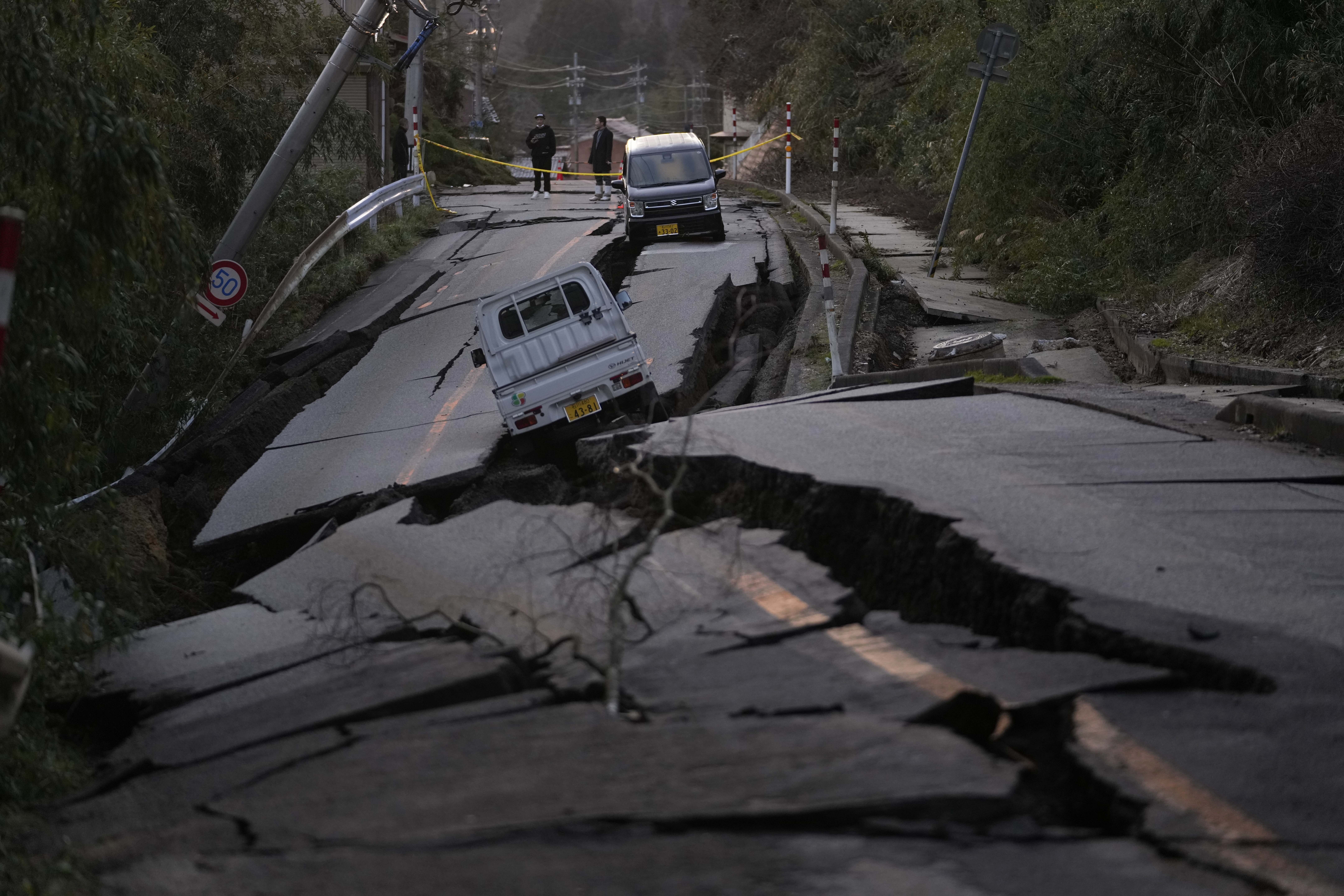 A road pushed up and ripped apart by the earthquake. There are huge cracks in the tarmac and two vans stuck inside. The road has been cordoned off behind yellow tape. Two people are looking at the scene.
