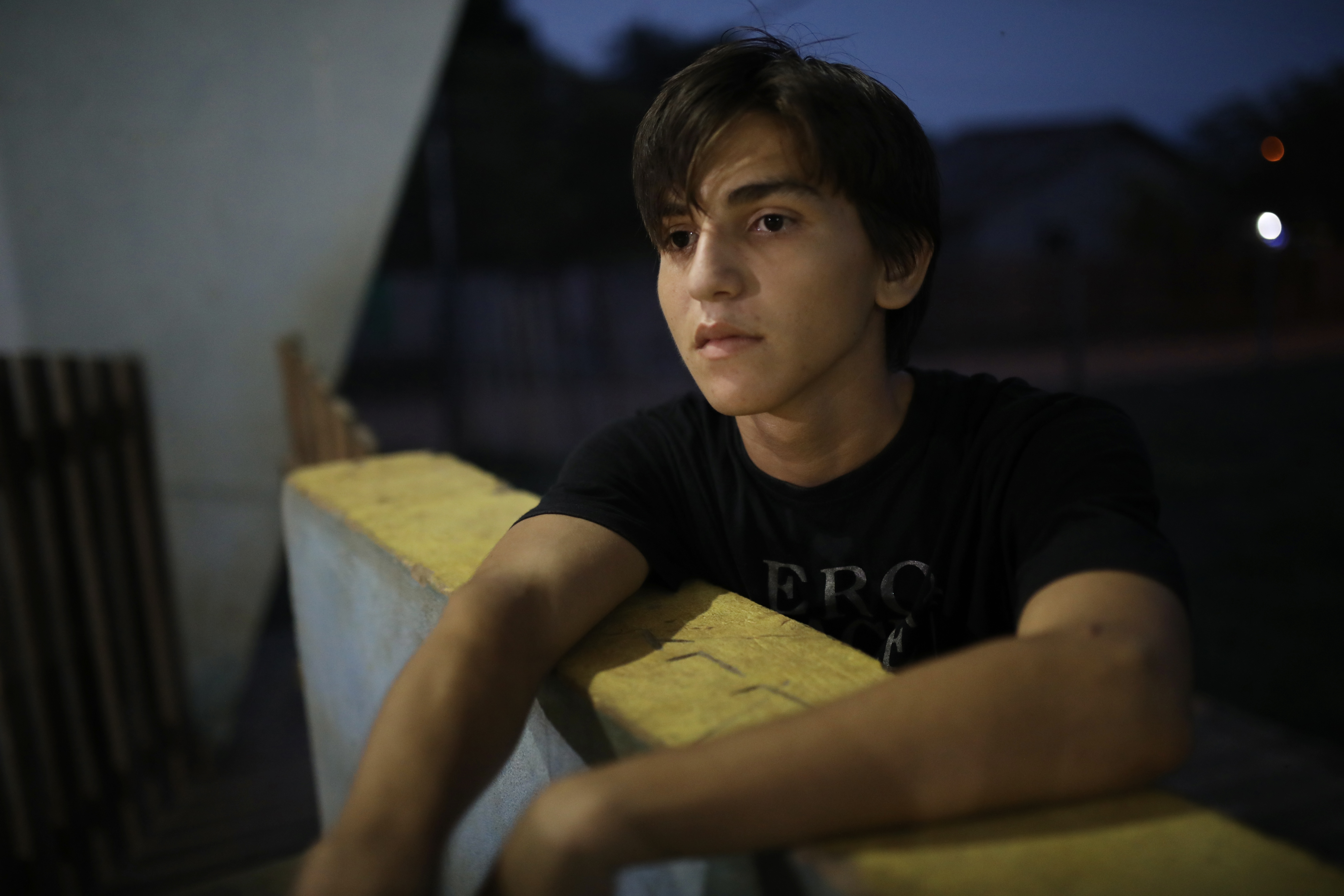 A teenage boy leans against a concrete wall. Behind him is a dark-blue nighttime sky.