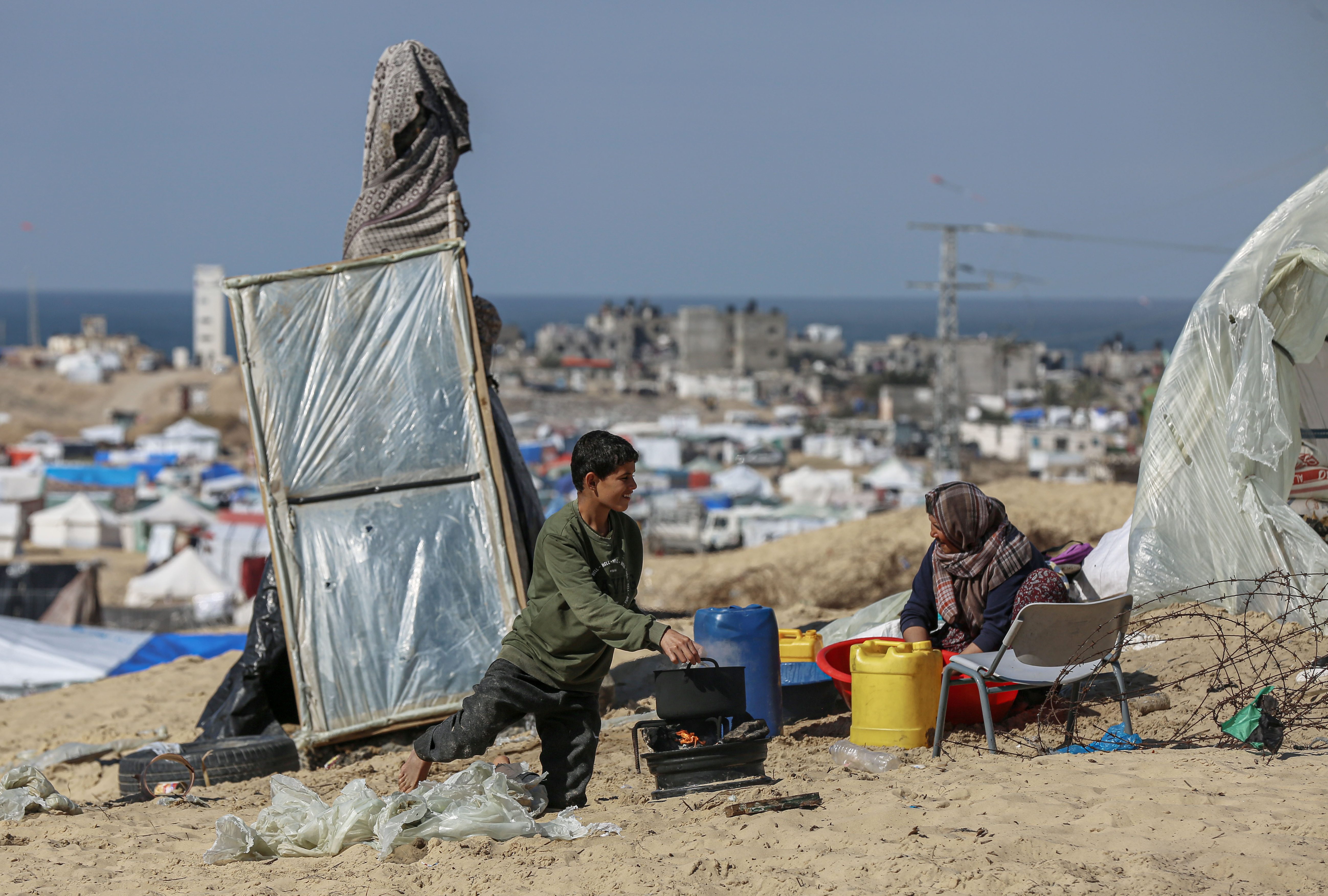 A boy is cooking on a makeshift fire, talking to a woman who is preparing something in a tub nearby. They are on the sandy ground, surrounded by barbed wire and makeshift shelters