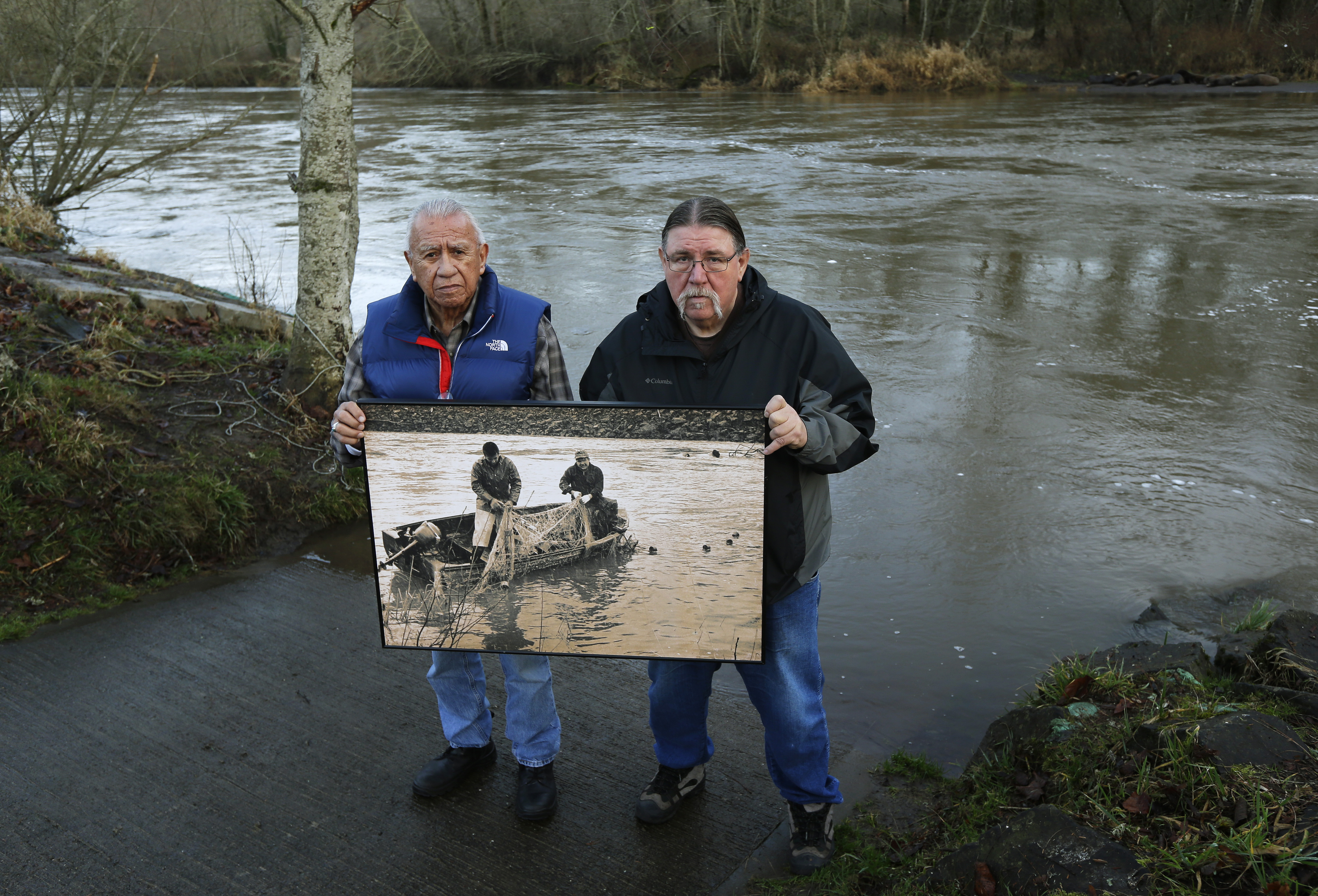 Billy Frank Jr and Ed Johnstone together hold an enlarged photo showing Indigenous ancestors fishing in the 1960s and 1970s where they are standing now.