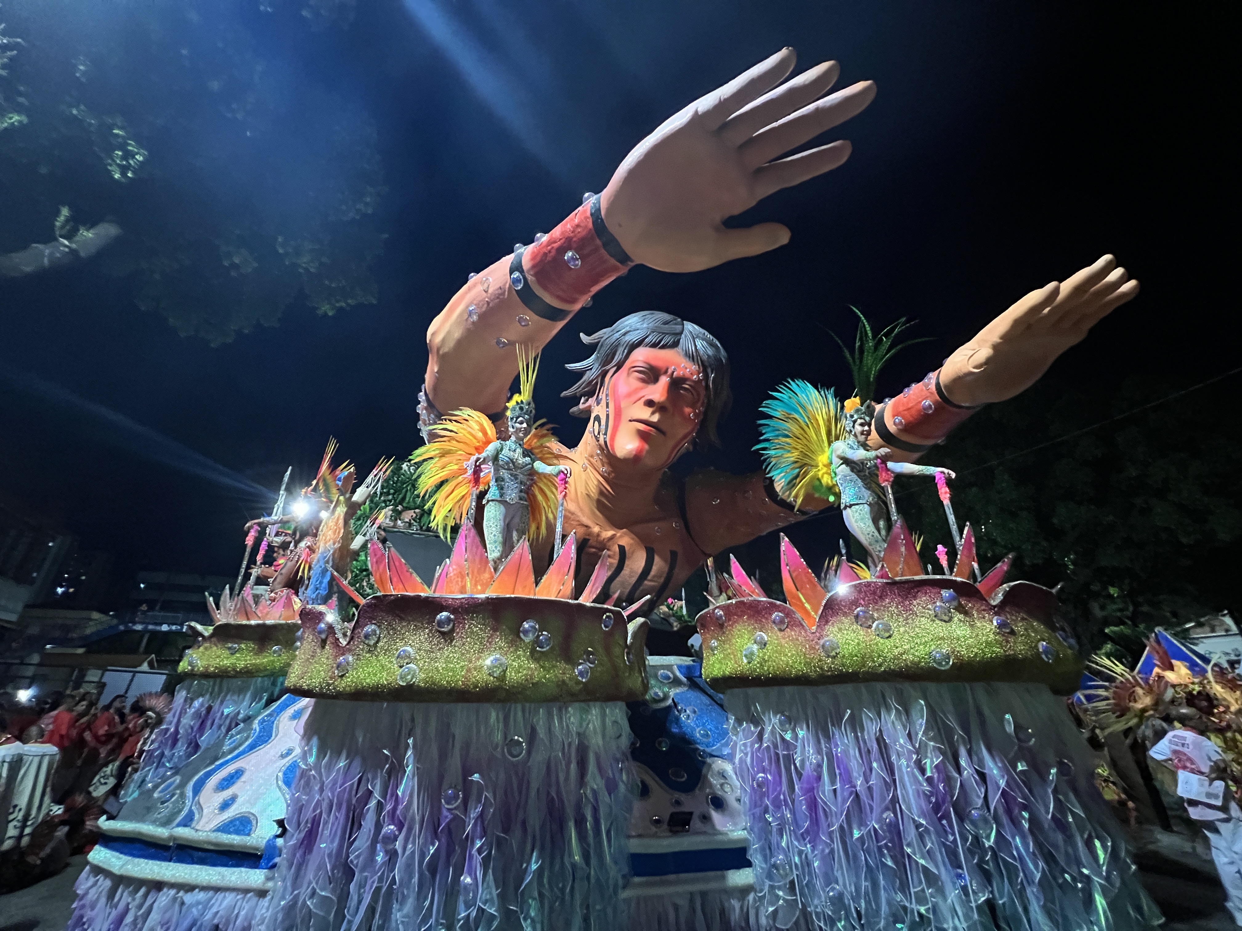 A float at the Sambadrome in Rio de Janeiro depicts a Yanomami person with his arms outstretched. On either side of him are platforms where dancers perform, dressed in blue sequins in a riot of yellow feathers emerging like wings from their backs.