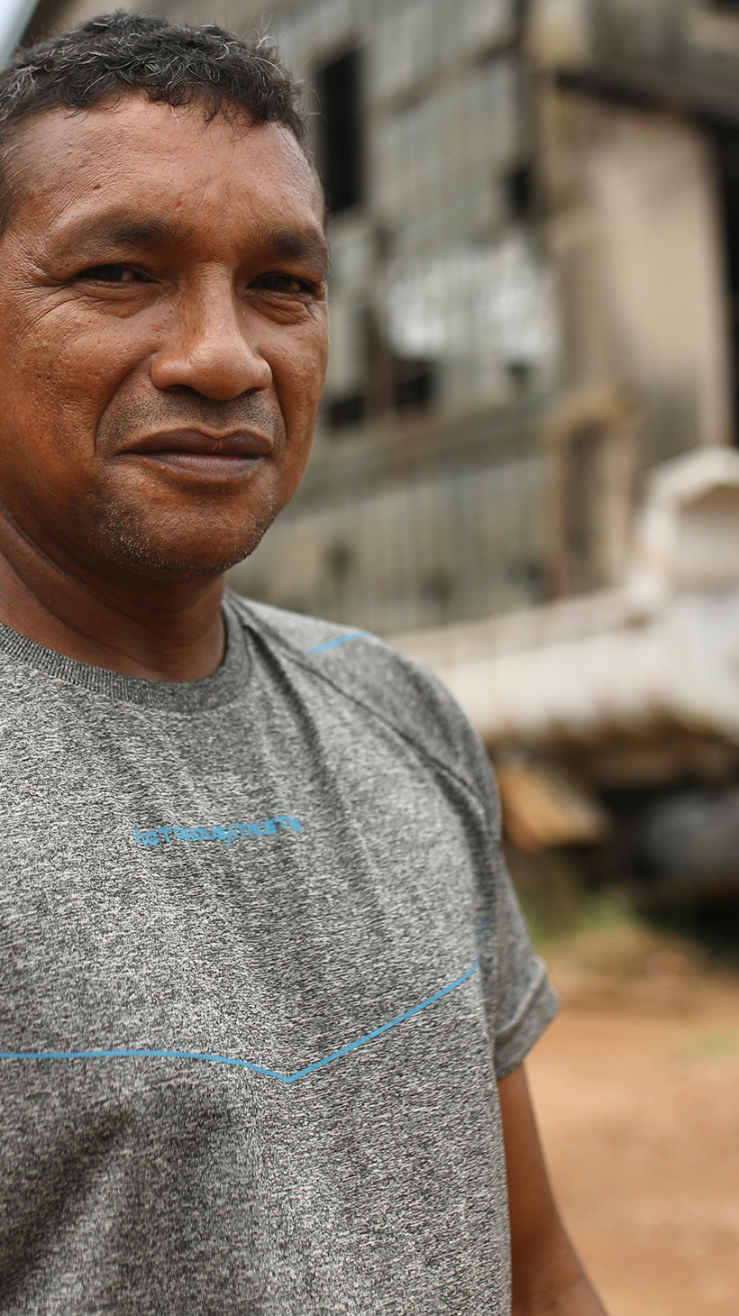 A man in a grey T-shirt stands in front of the abandoned warehouses at Fordlandia, an industrial town in the Amazon. A water tower is also visible behind him.