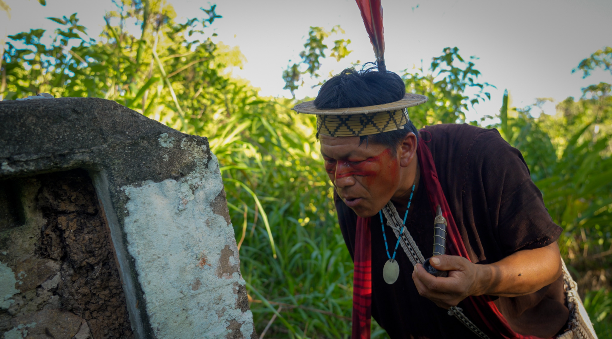 Ashaninka leader Tsitsiri Samaniego — dressed in a tunic and a straw headdress with three macaw feathers sticking upwards — inhales from a tobacco pipe as he stands before a headstone for his grandfather.