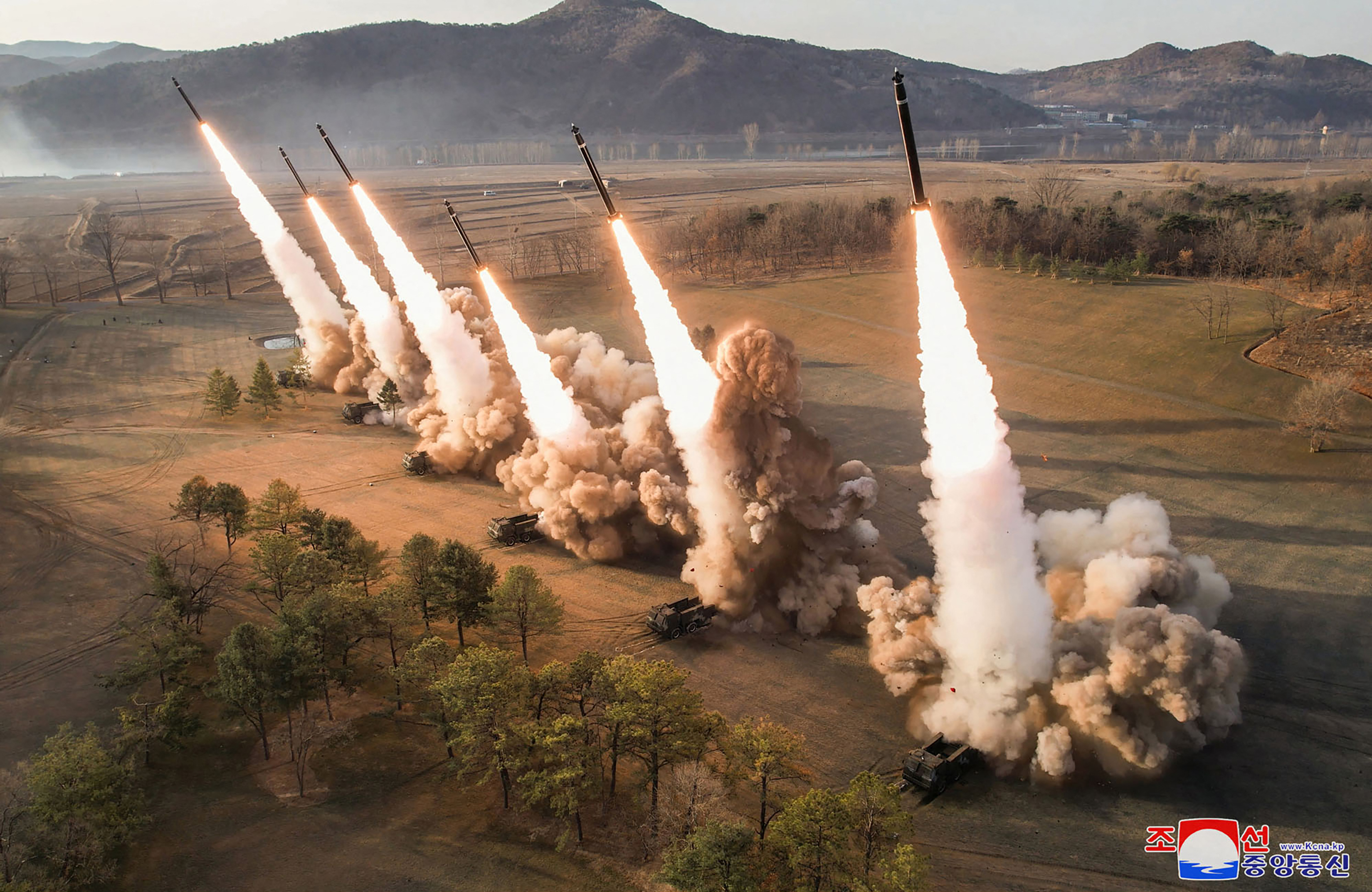 Rockets launching into air from a row of launchers. There's a lot of smoke and flame. There are mountains behind.