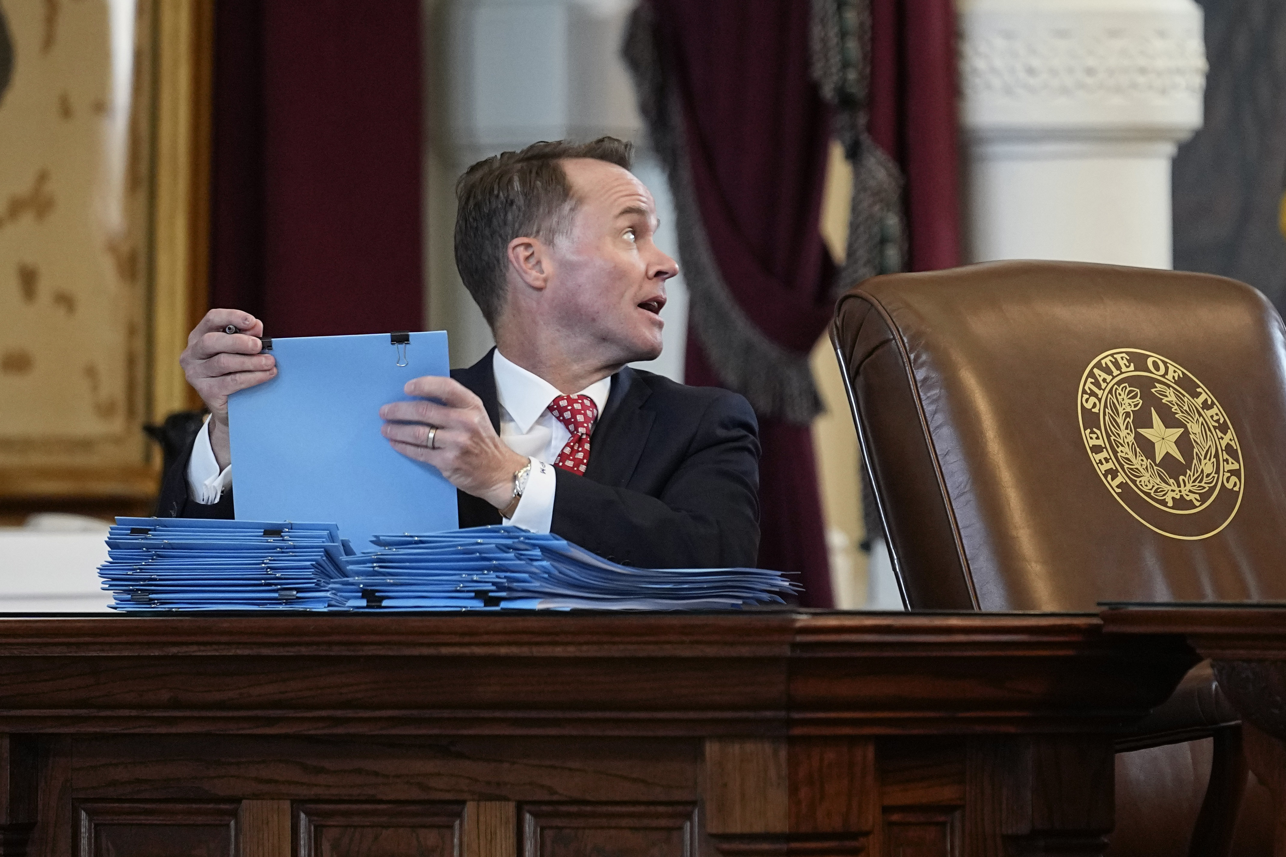 Dade Phelan sits behind a large wooden desk in the legislature, with a pile of blue folders before him.