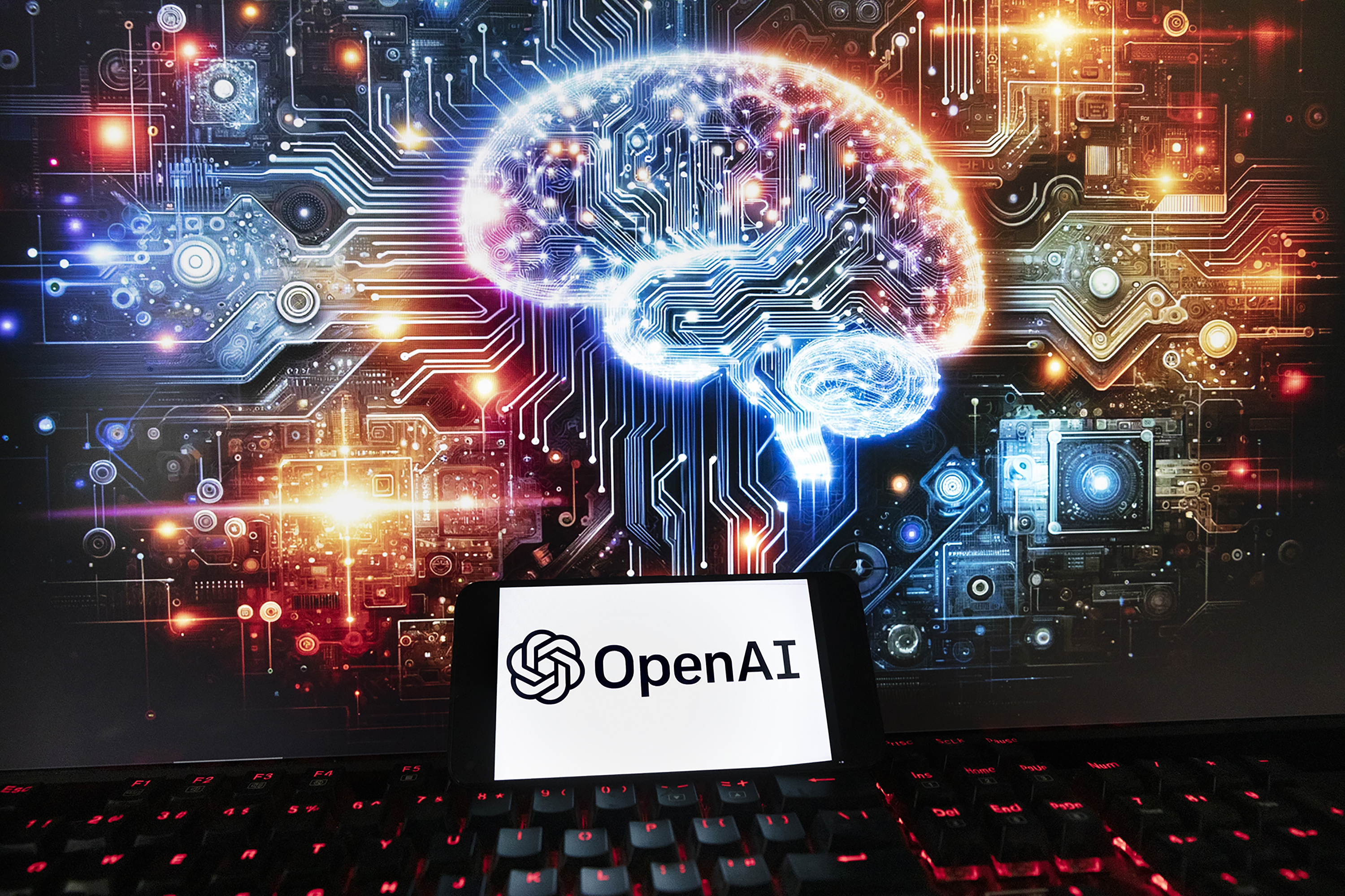 The OpenAI logo is displayed on a cell phone with an image on a computer monitor