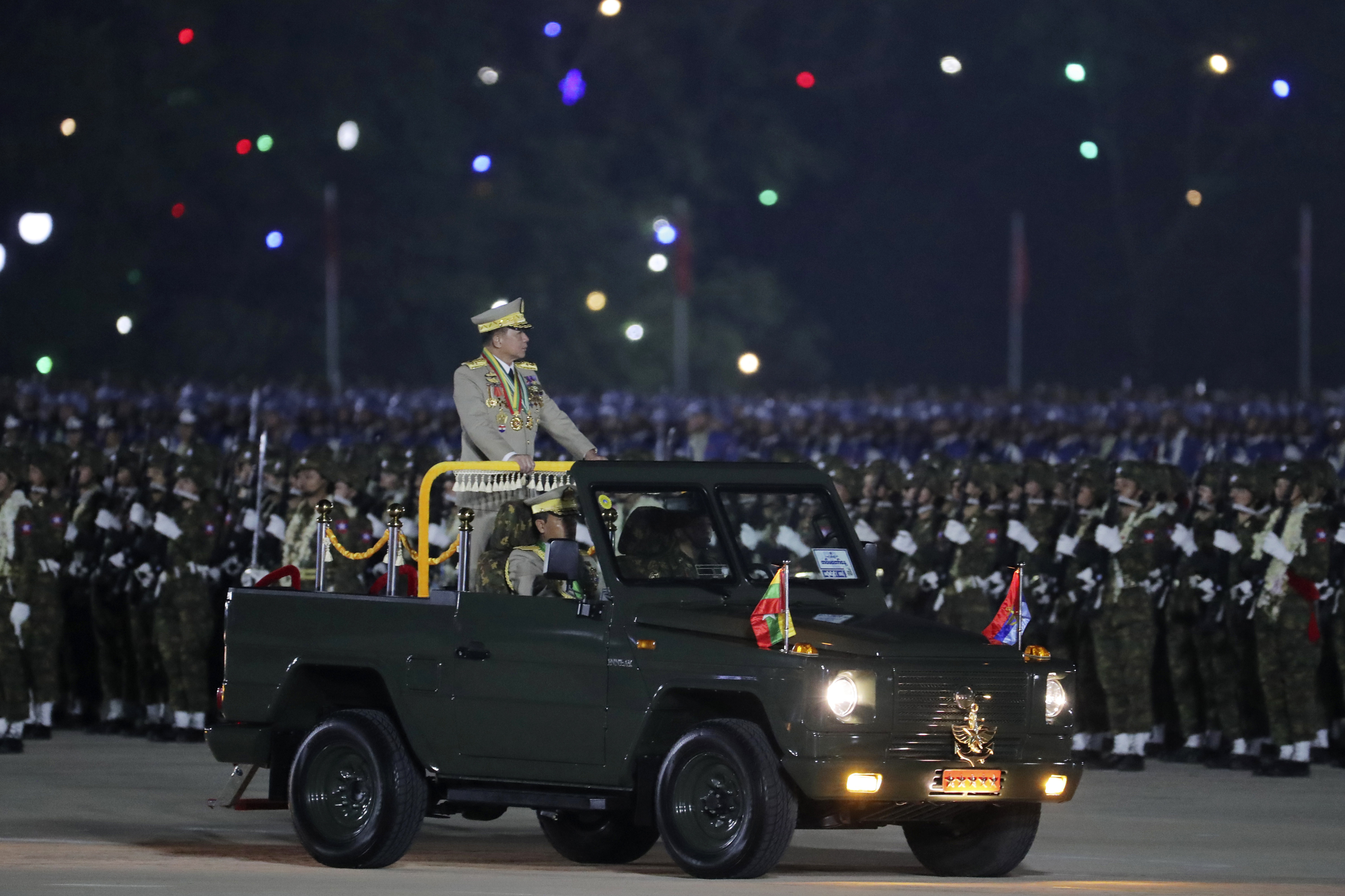 Min Aung Hlaing inspecting troops at the annual Armed Forces Day parade. His is standing on the back of an open-topped jeep-like vehicle. It is night time.