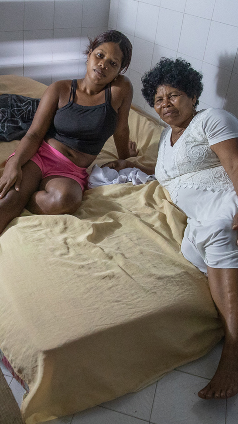 Two women lie on a mattress plopped on the floor of a coliseum.