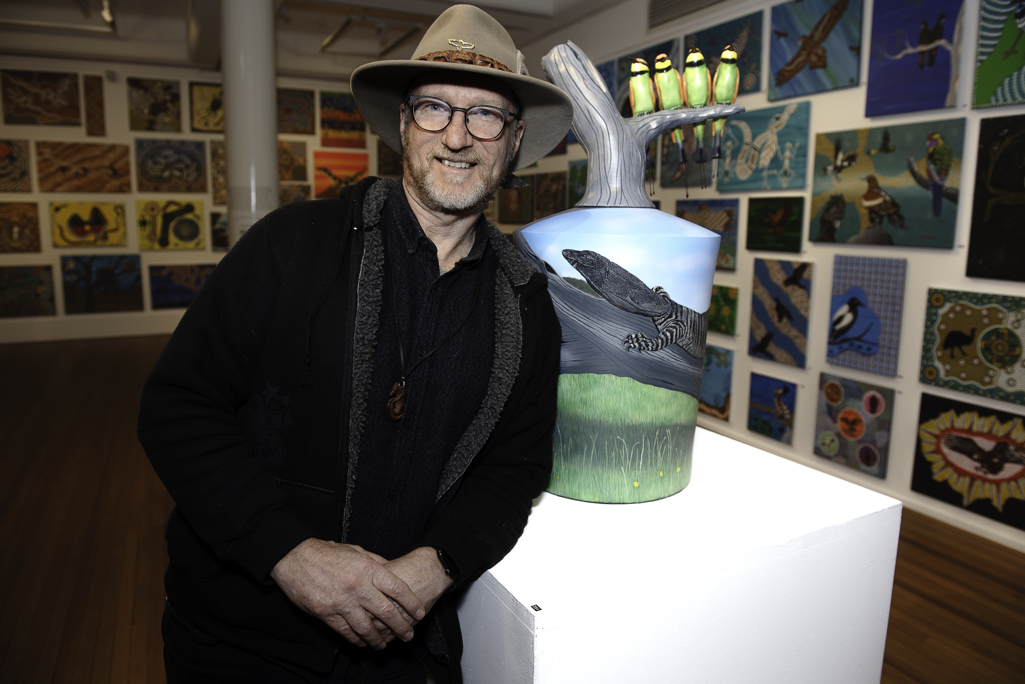 Sean Miller. He is standing in front of a ceramic art work. He's wearing a black short and coat as well as a hat.