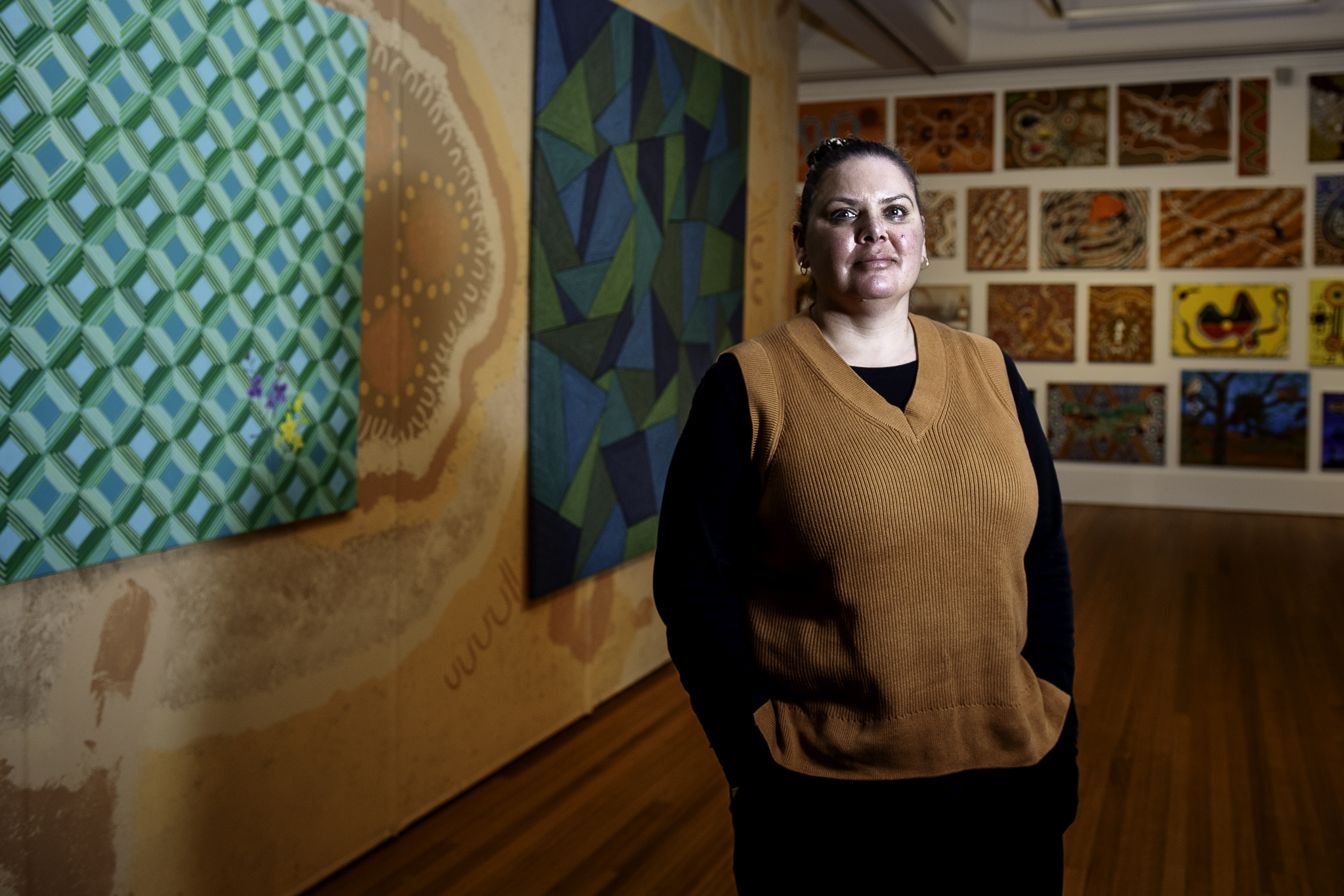 Stacey Edwards. She is standing in front of her art work. She is wearing a black shirt with a brown vest over the top. Her hair is tied back and she has her hands in her pockets