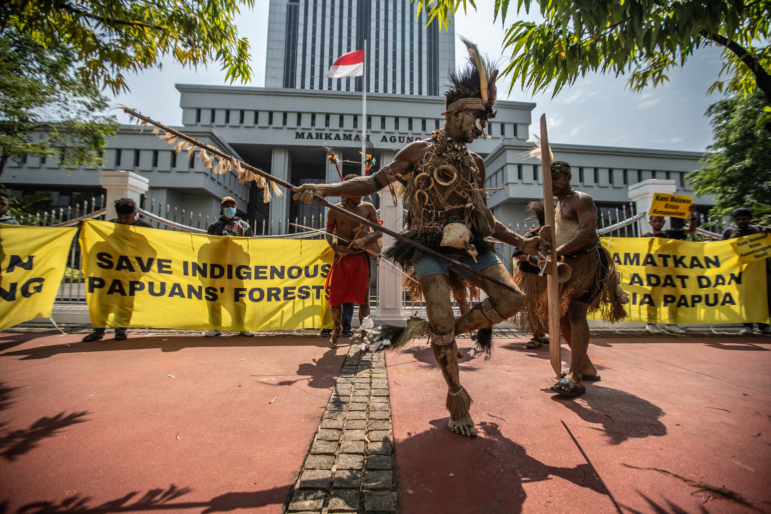 Representatives of the Awyu and Moi Indigenous Peoples from West Papua dance outside the Supreme Court gates. They are wearing traditional clothing. There is an Indonesian flag hanging from the court building.