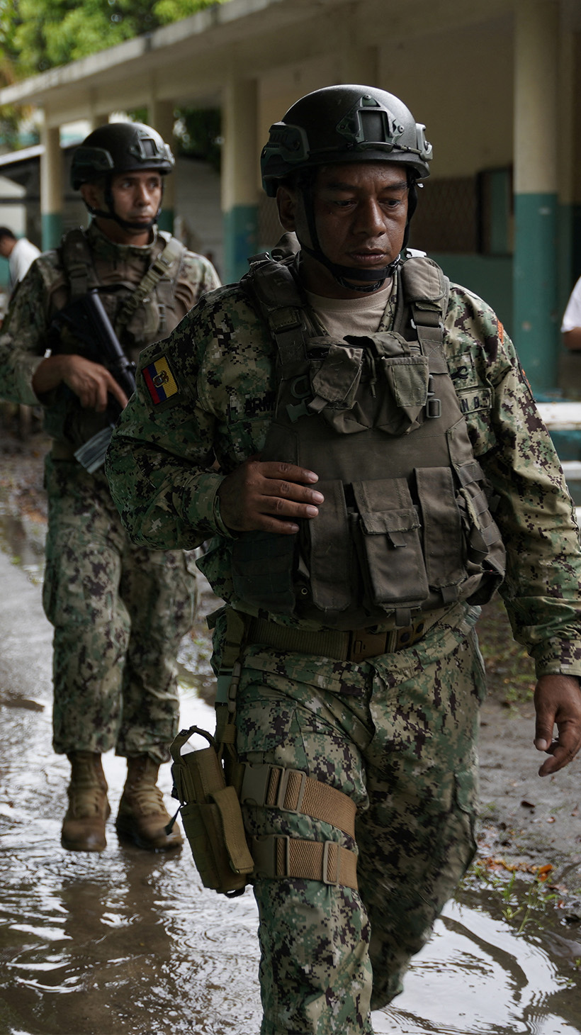 Two soldiers in helmets and fatigues walk in a row down a muddy path between buildings in Guayaquil.
