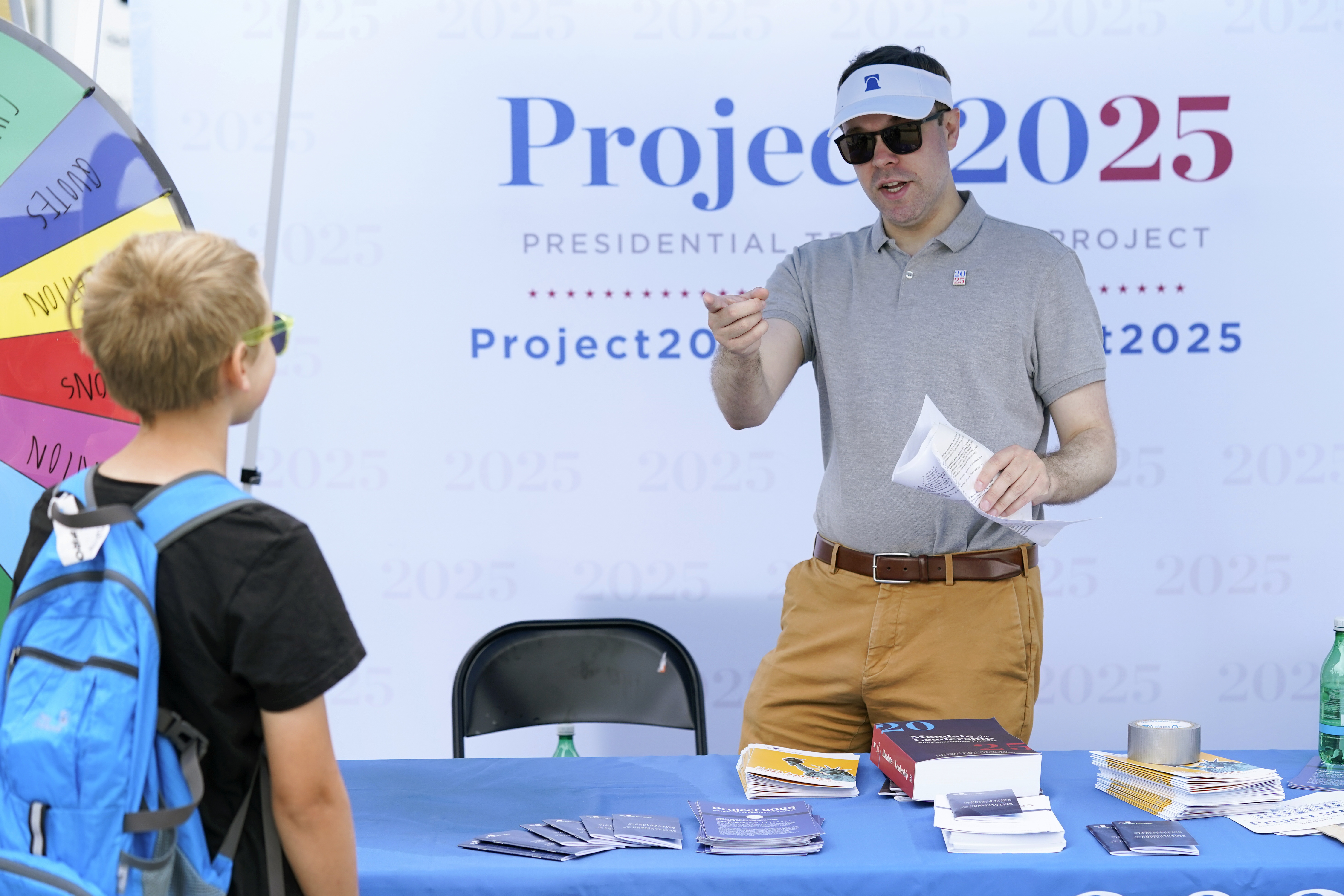 A man in a visor and a polo shirt stands behind a table under an outdoor tent and speaks to a child. Behind him is a sign that reads, "Project 2025."