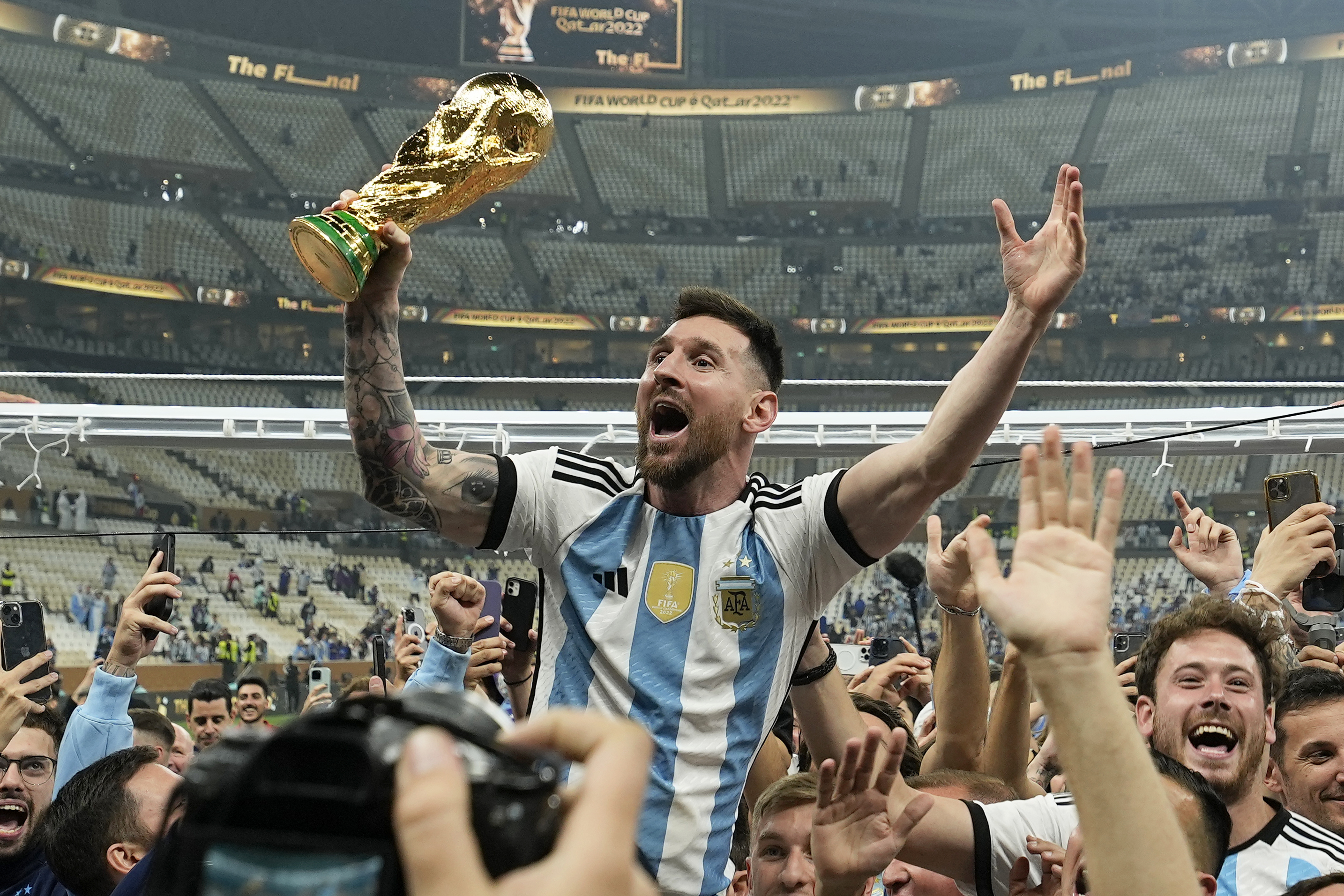 Lionel Messi, dressed in his blue and white Argentine stripes, is raised aloft by a crowd of supporters. In one hand, he carries the FIFA trophy.