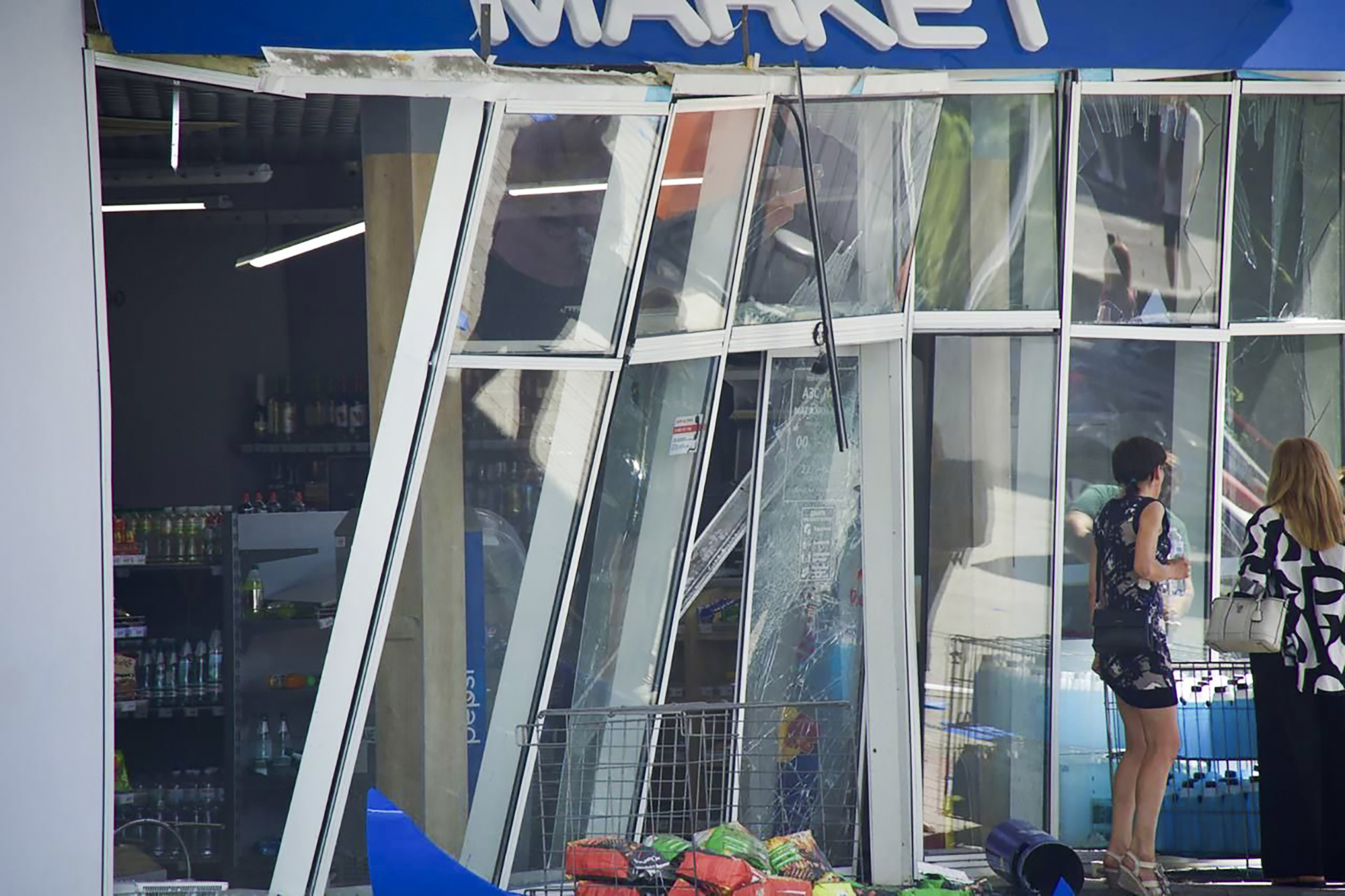 A damaged shop facade after a Russian attack on Dnipro. The window and door frames are bent and sticking outwards. Some of the glass has broken. Two women are standing outside.