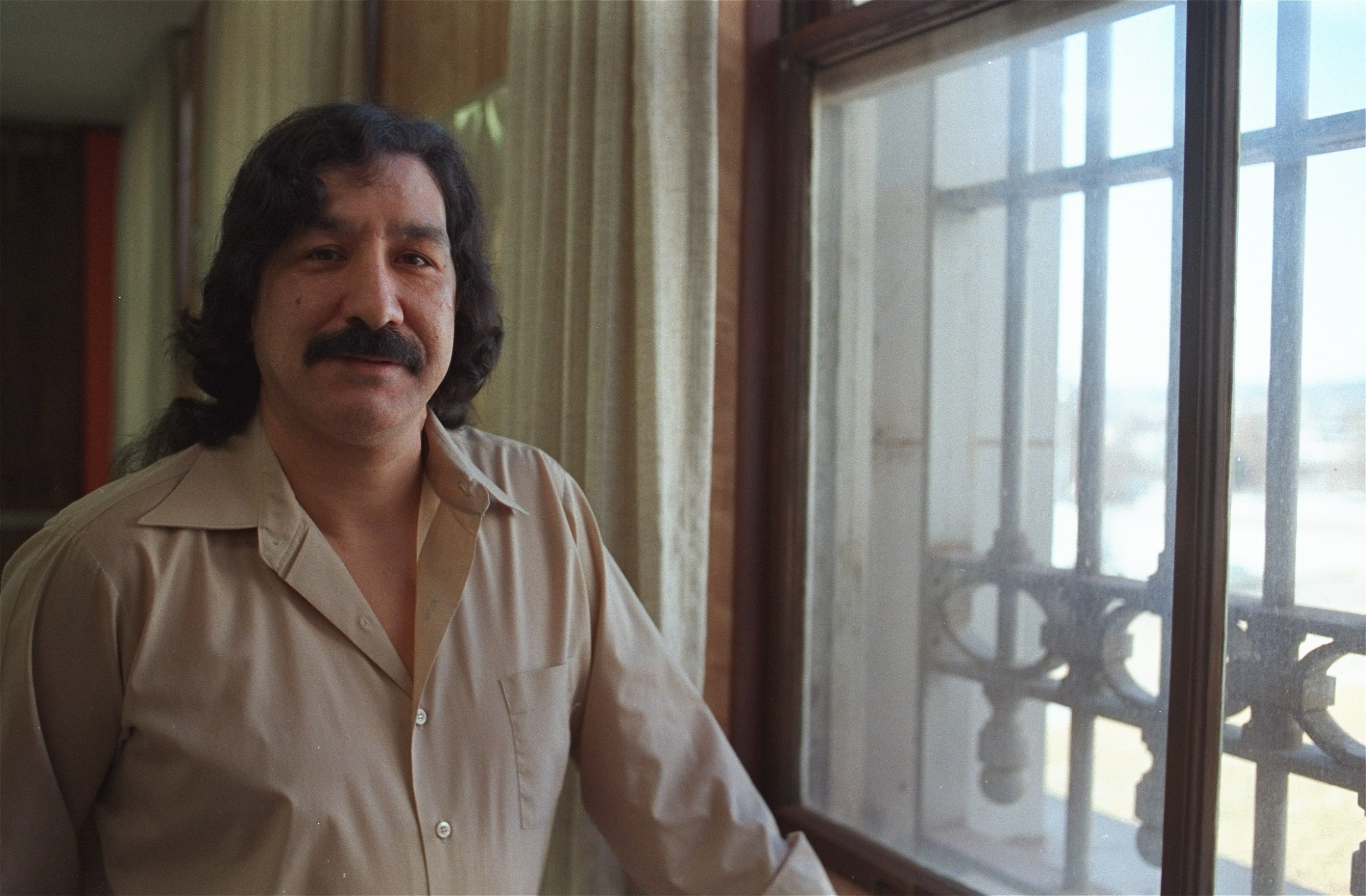 Leonard Peltier stands at a prison window, dressed in a beige collared shirt.