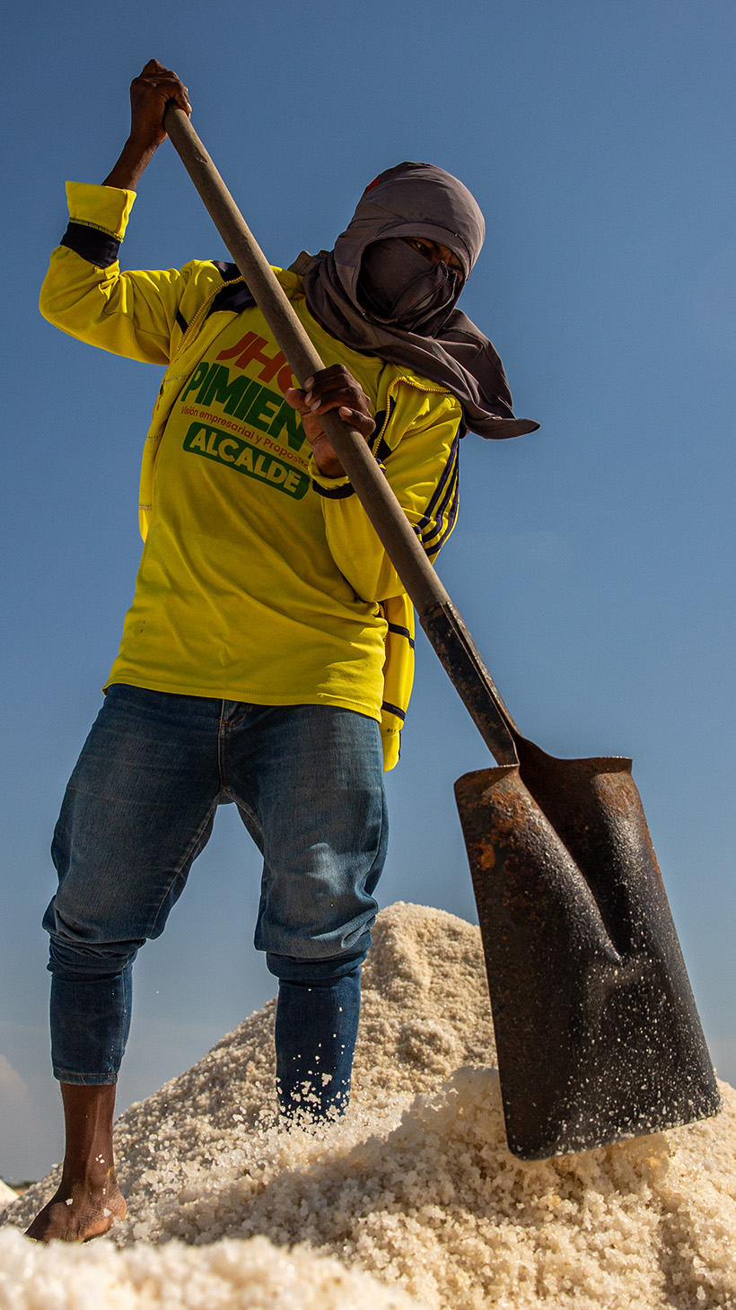 A man — his head wrapped in a scarf to protect against the wind and heat — uses a shovel to pile up salt.