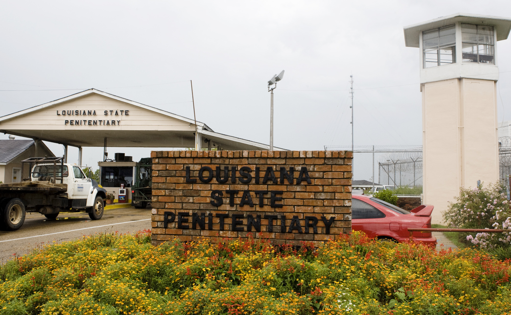 A view of the entrance of the Louisiana State Penitentiary in Angola. A watch tower rises on one side, next to a covered entrance way with a security checkpoint that vehicles pass through. In the front sits a brick wall with the words: Louisiana State Penitentiary.