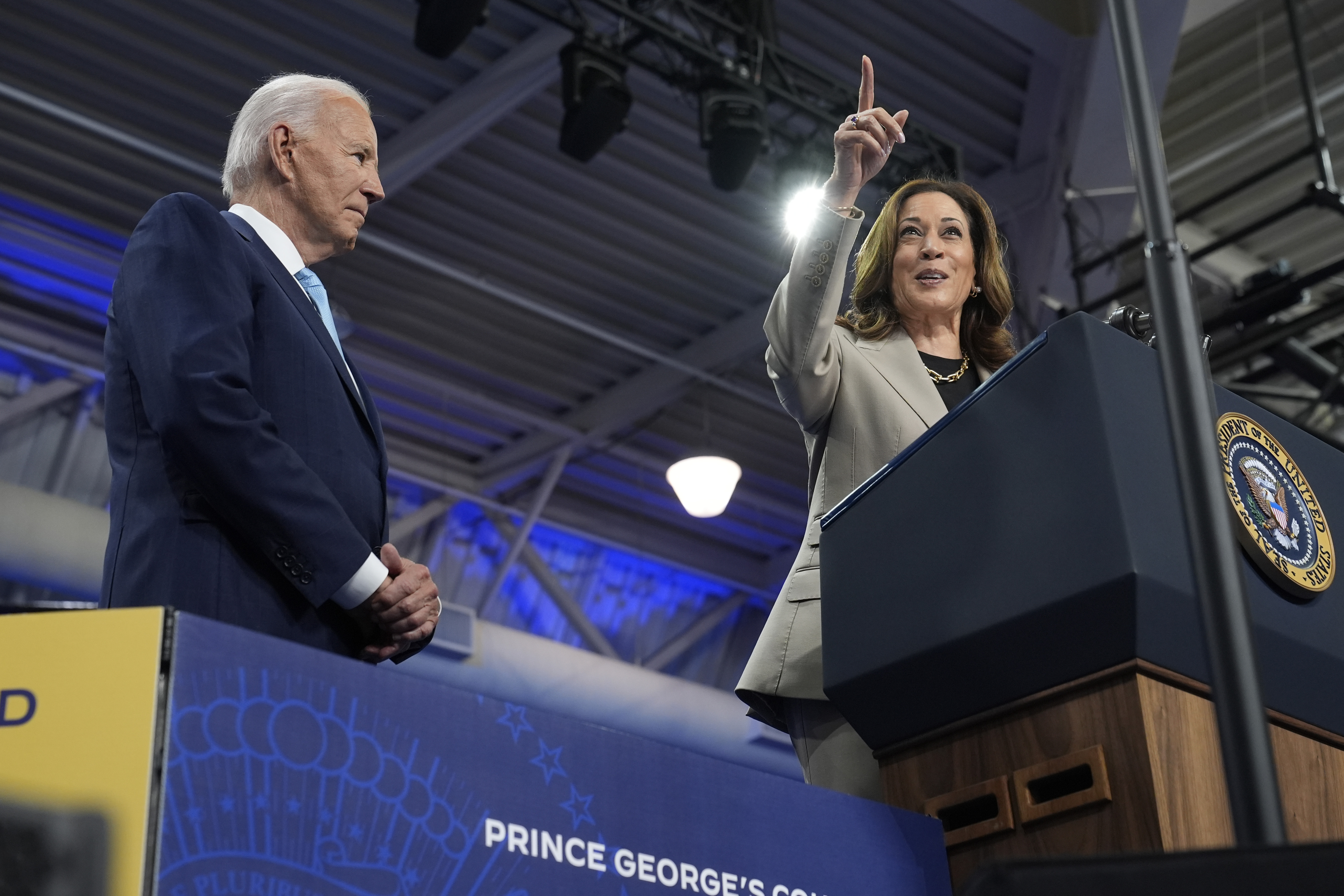 Kamala Harris stands at a podium pointing, while President Joe Biden looks on.