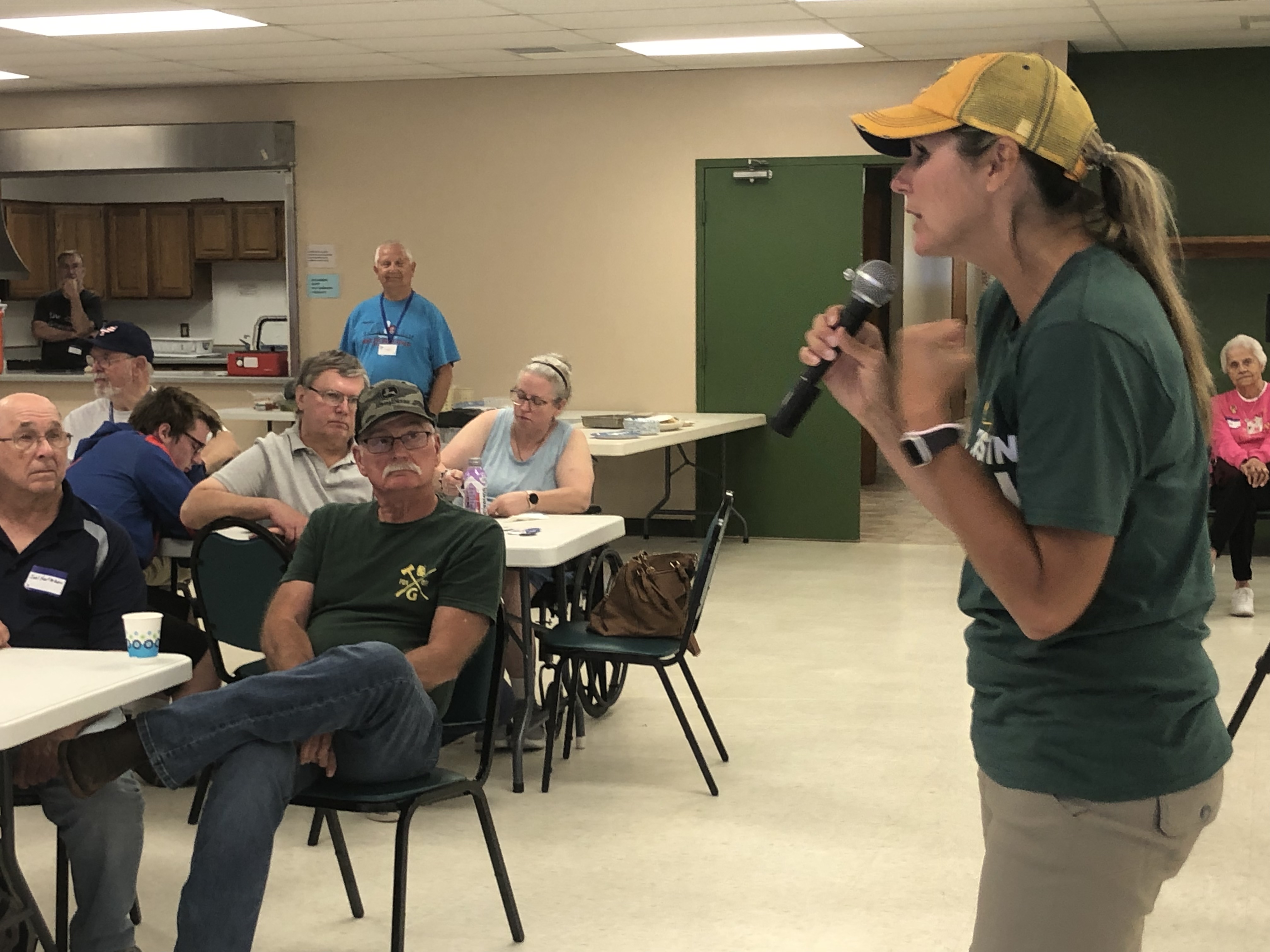 Kristin Lyerly, dressed in a yellow baseball cap and green shirt, speaks into a microphone in front of a room of voters.