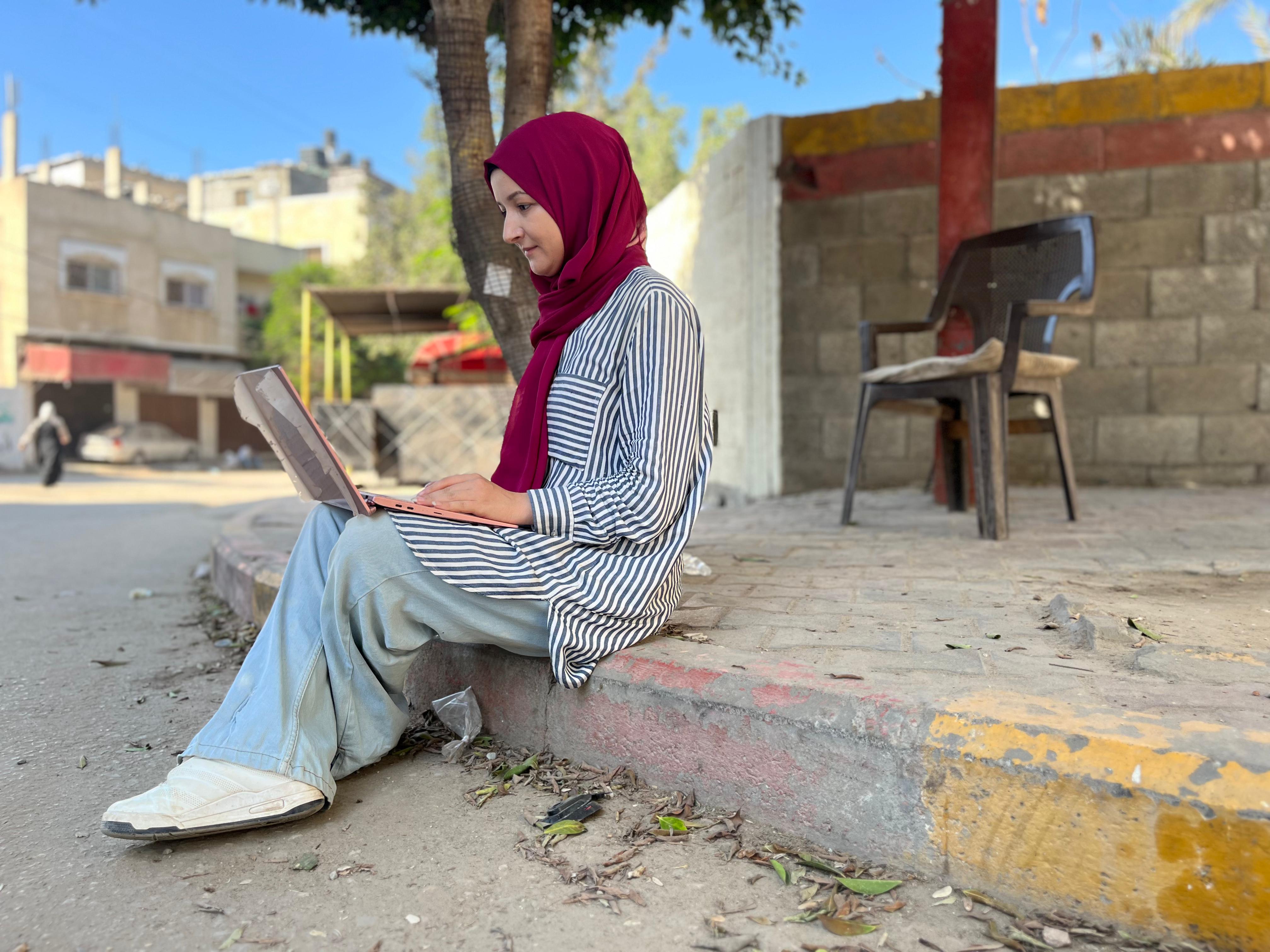 Maha Hussaini in the striped shirt she threw on when she left her house some 10 months ago. She is sitting on a sidewalk in Gaza working on her laptop