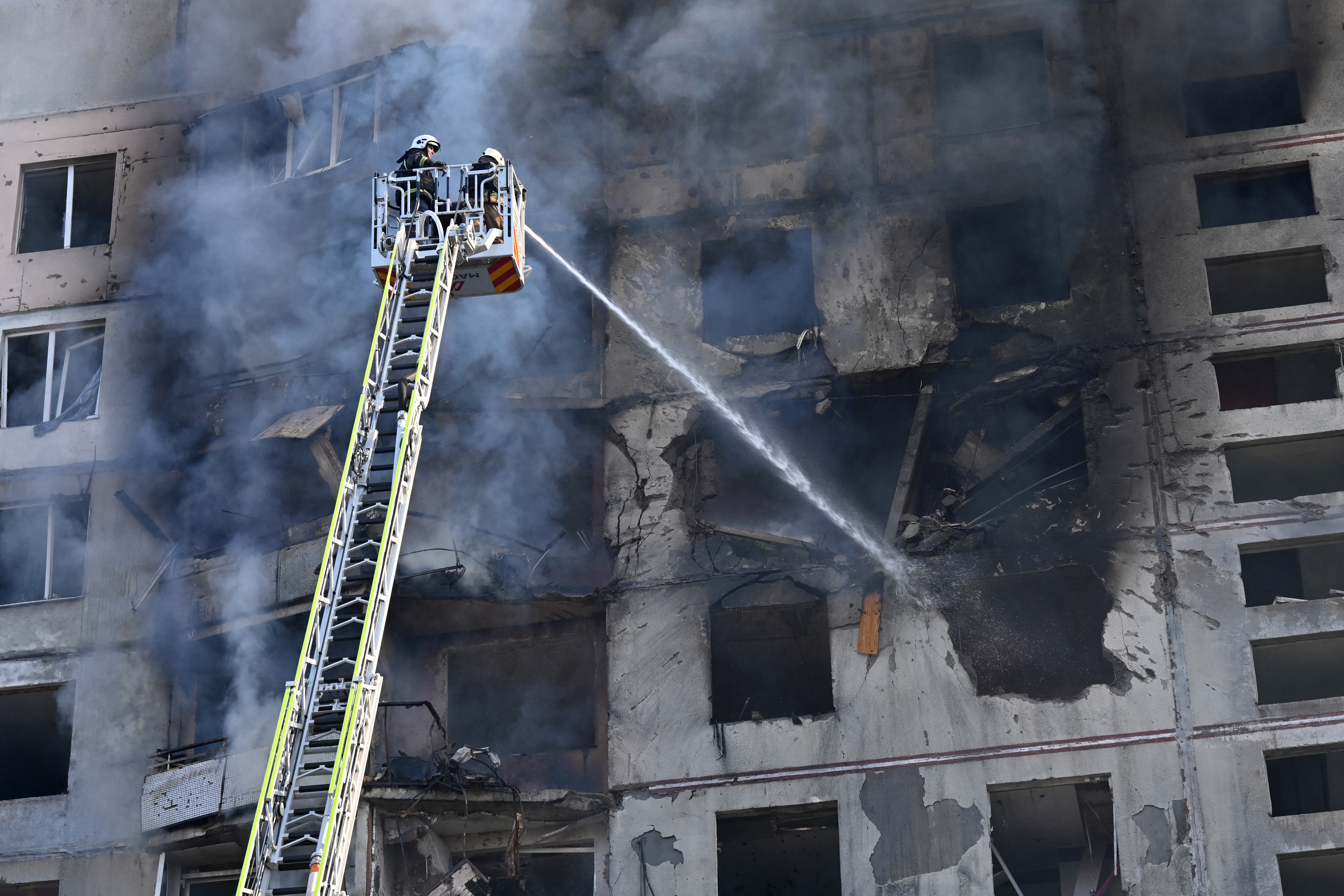 A firefighter on an extended ladder targeting a fire at a high rise apartment blocl. The walls of the building are black from the fire. Smoke is billowing from the building.