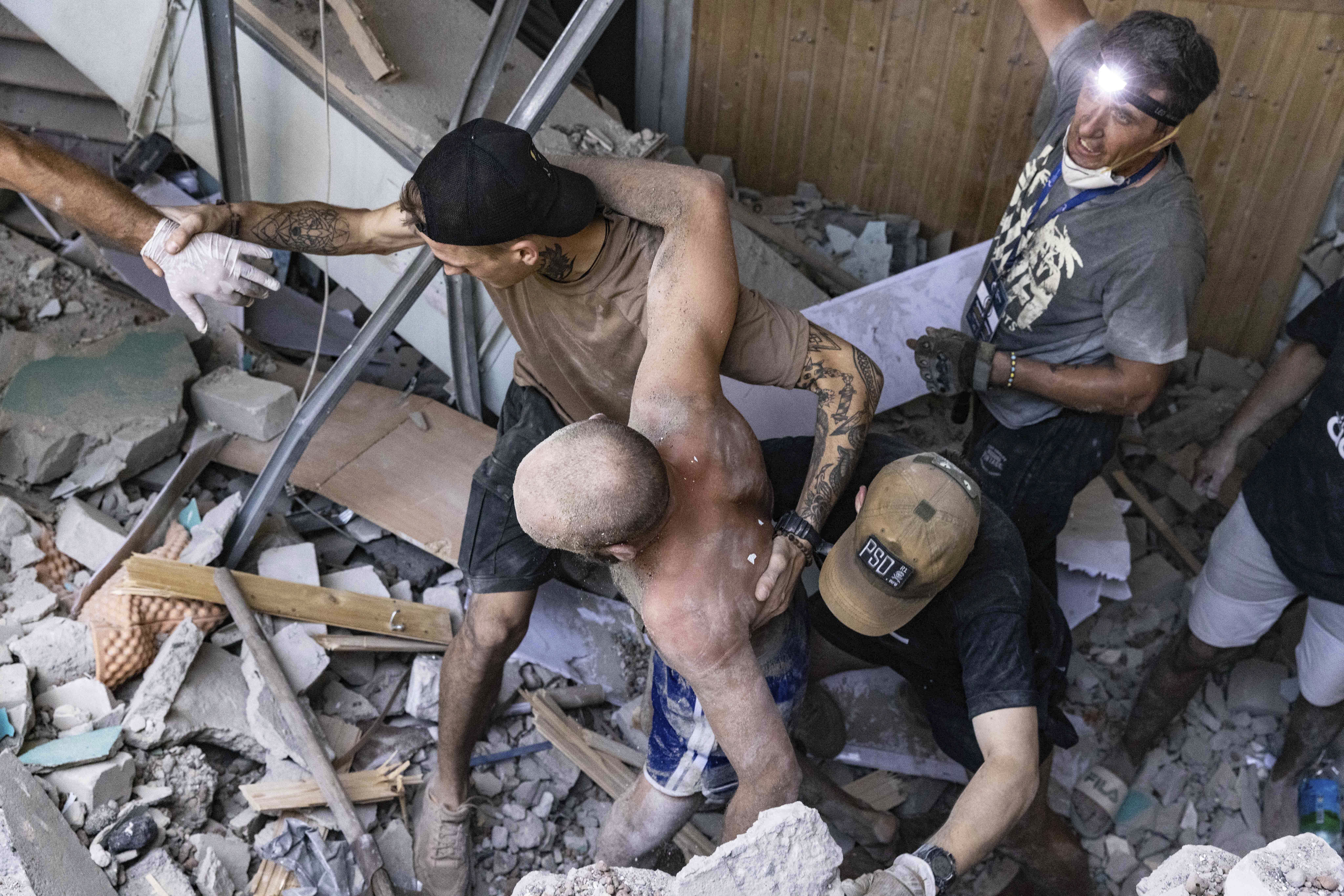 A man without a shirt and covered in dust making his way out of a ruined building. He is injured. Other people are helping him.