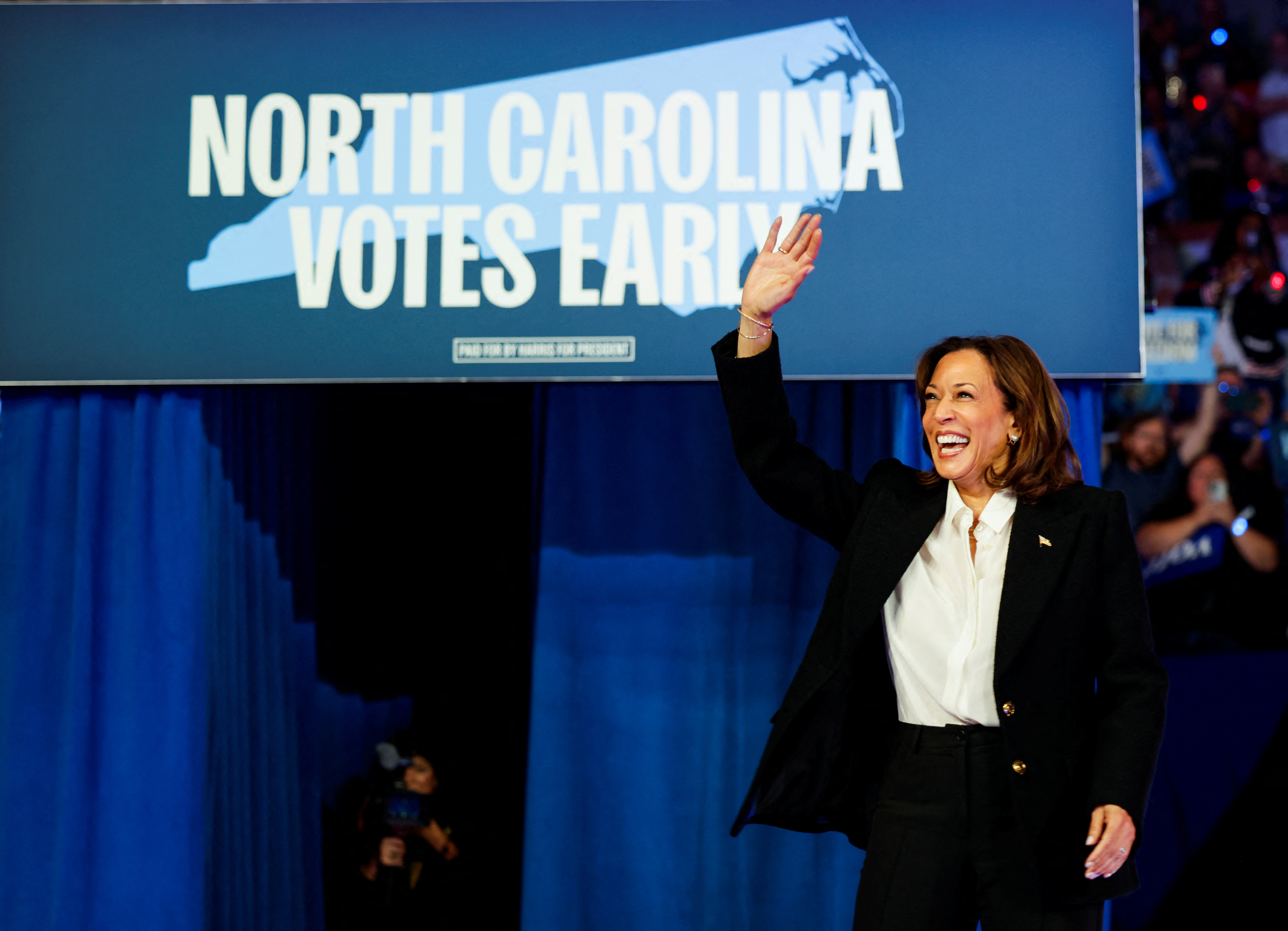 Kamala Harris waves in front of a sign that shows an outline of North Carolina. It reads: