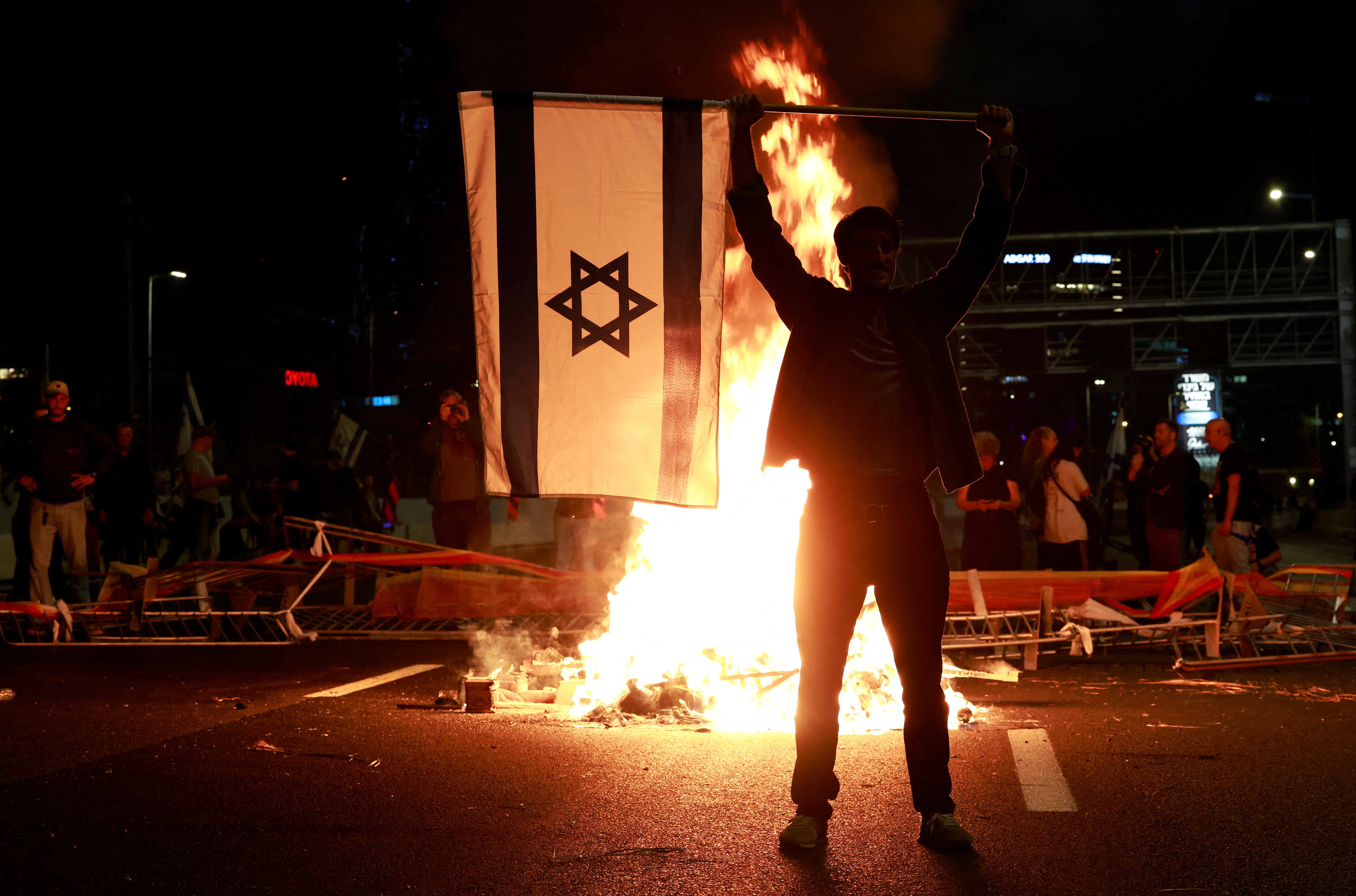 Silhouette of protester standing in front of lit fire at nighttime and holding an Israeli flag