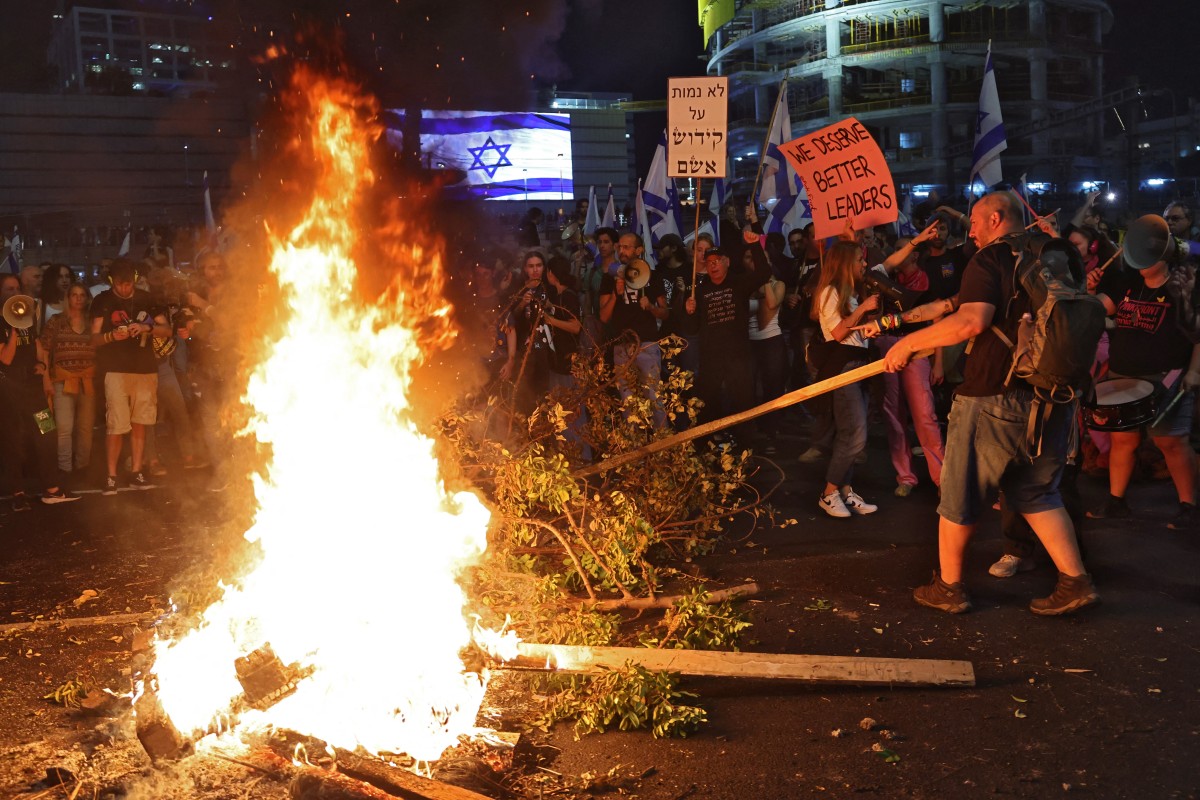 A man puts large tree branches into a fire with a group of people in the background holding placards. One says, "We need better leaders."