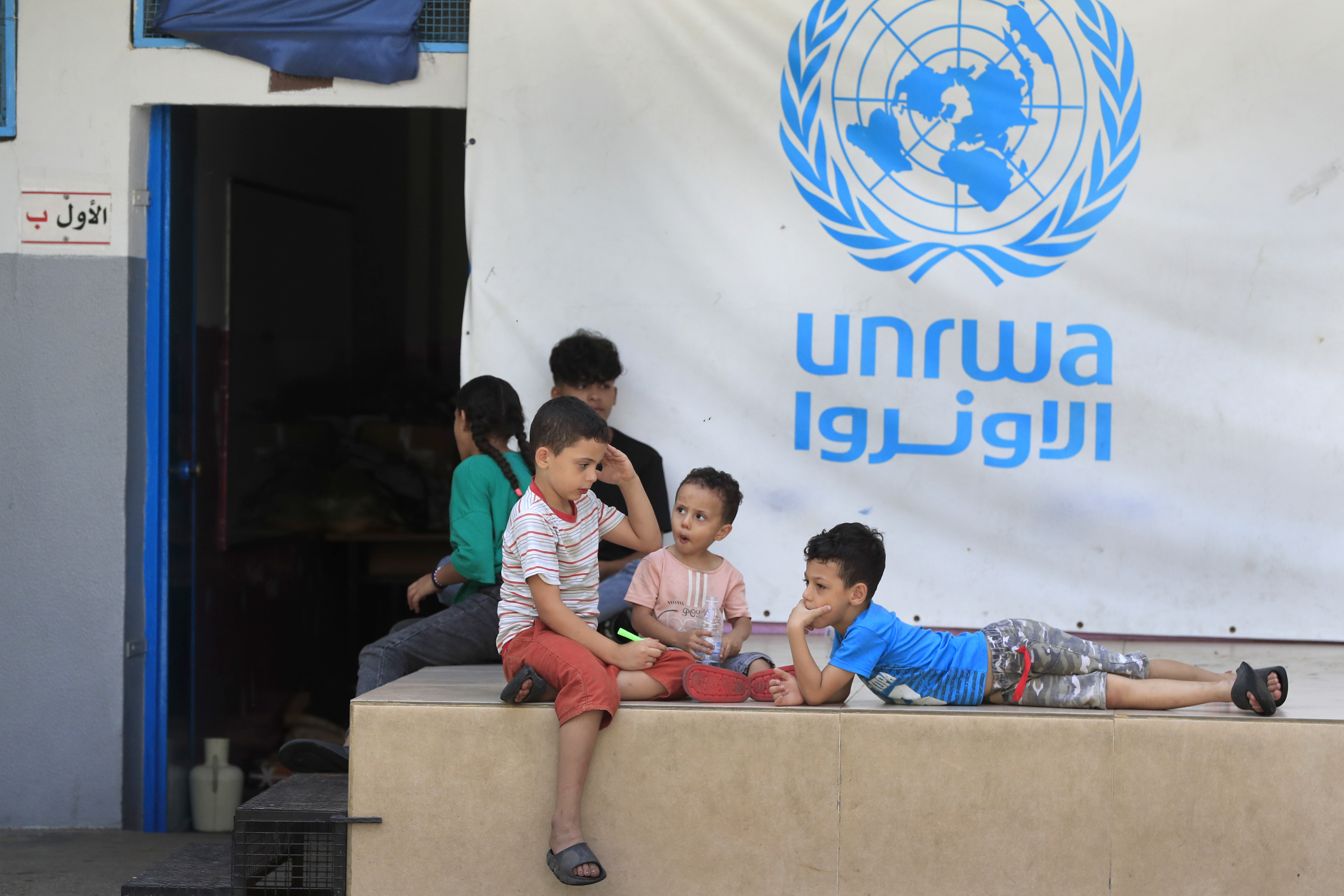 children sit next to a blue sign that says UNRWA
