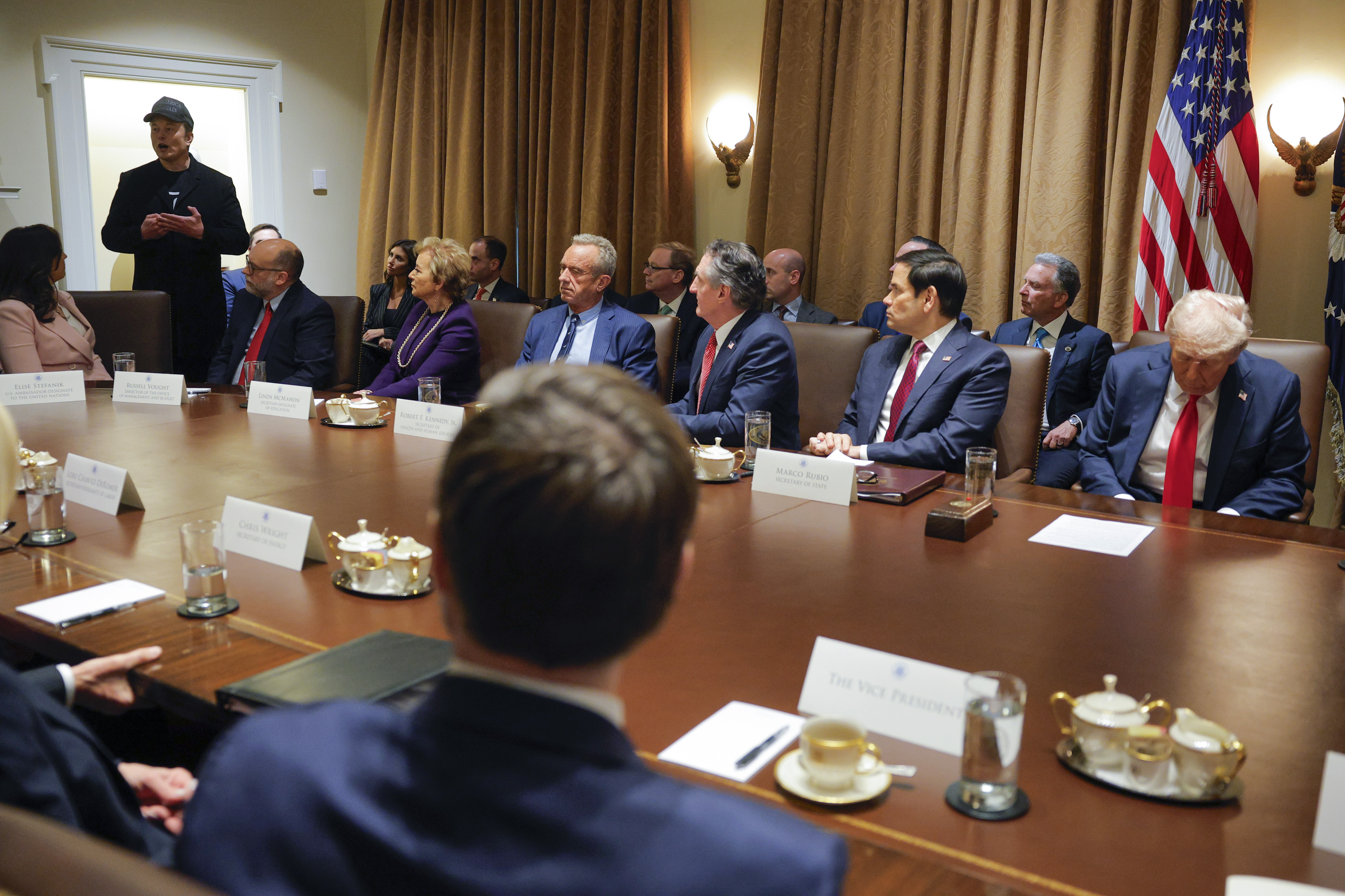 Elon Musk stands at the end of the cabinet table, speaking.