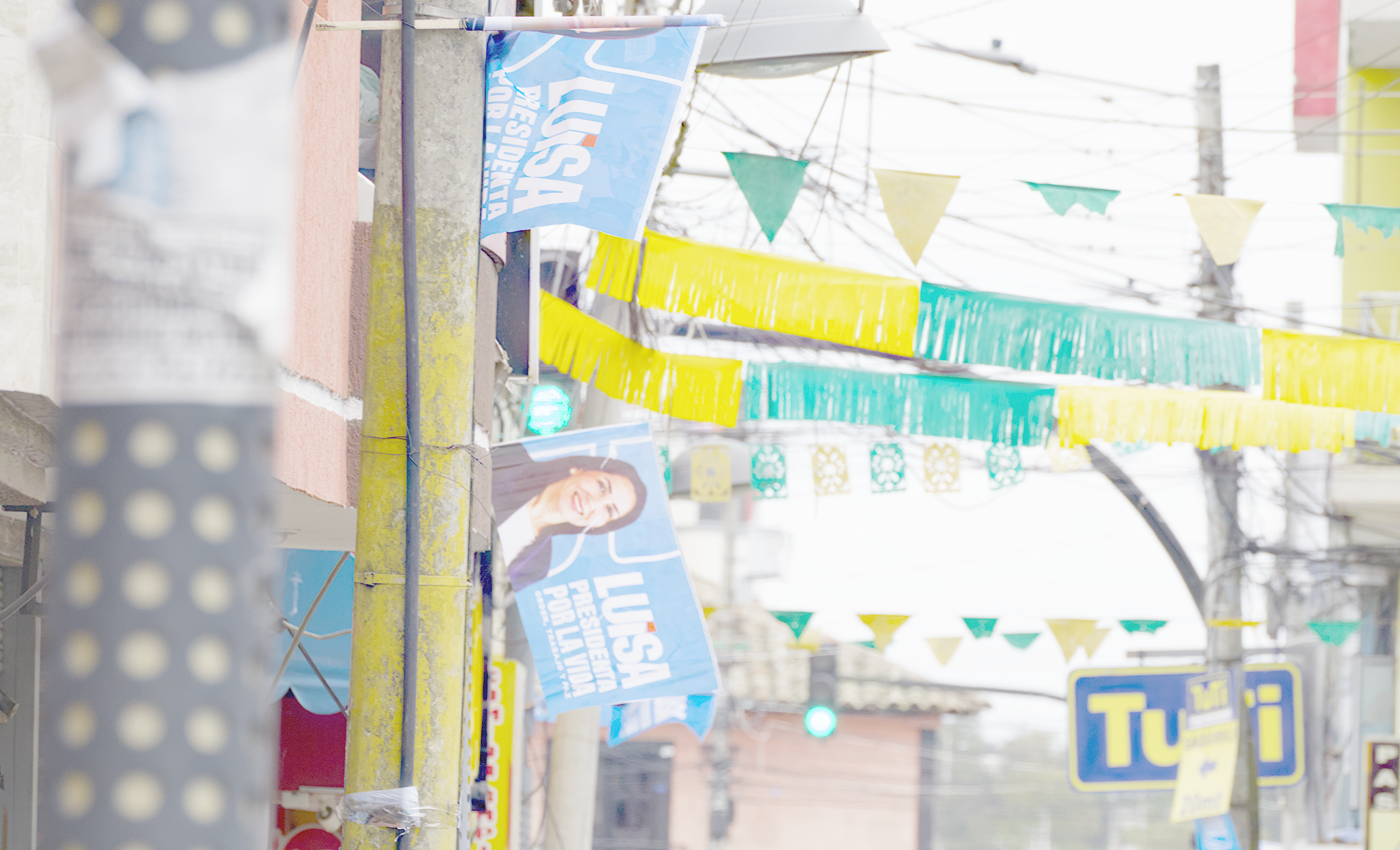 Flags for Luisa Gonzalez's presidential campaign fly above an Ecuadorian street decorated with yellow and green flags and banners.
