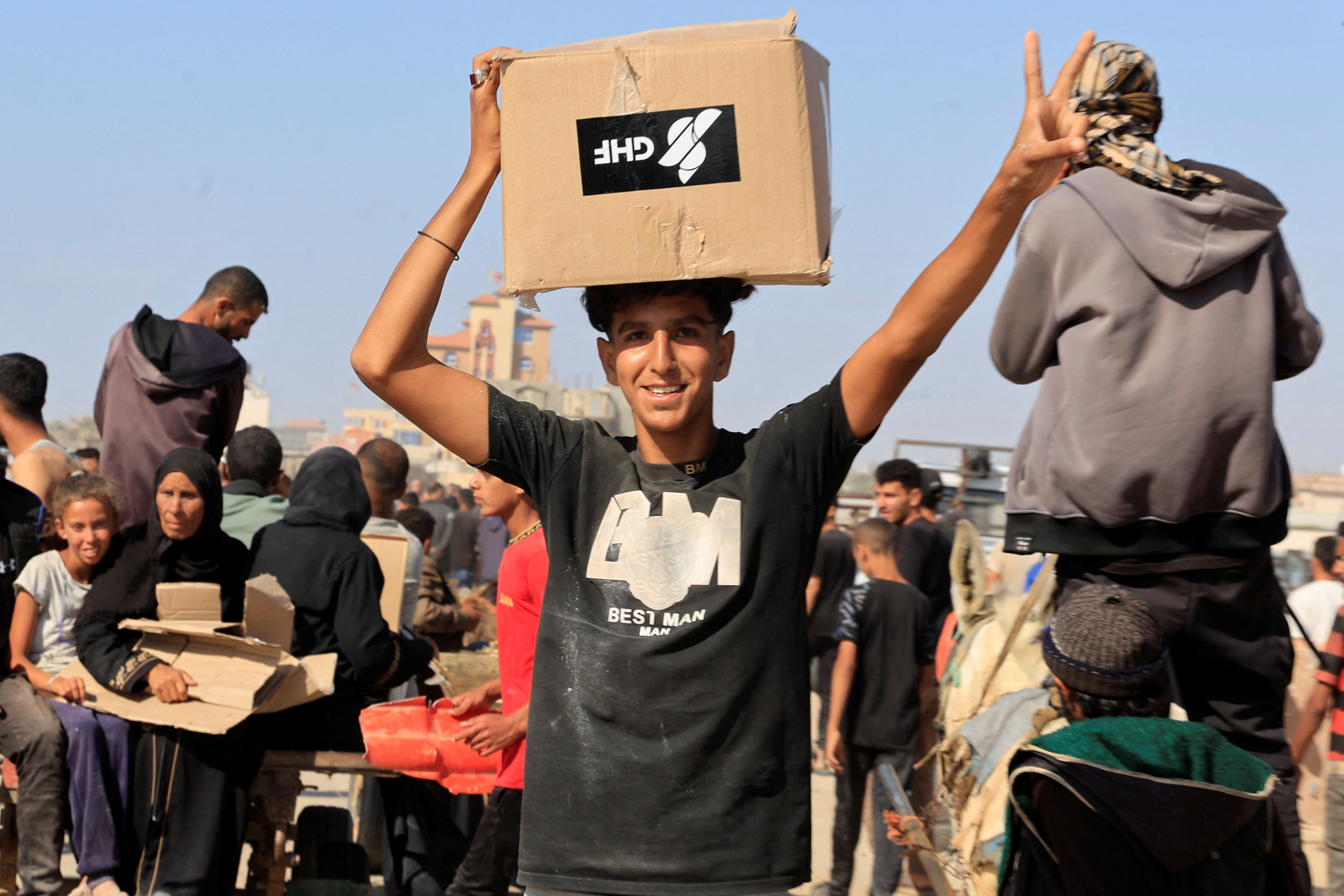 A boy flashes a peace sign as he balances a cardboard box of supplies on his head.