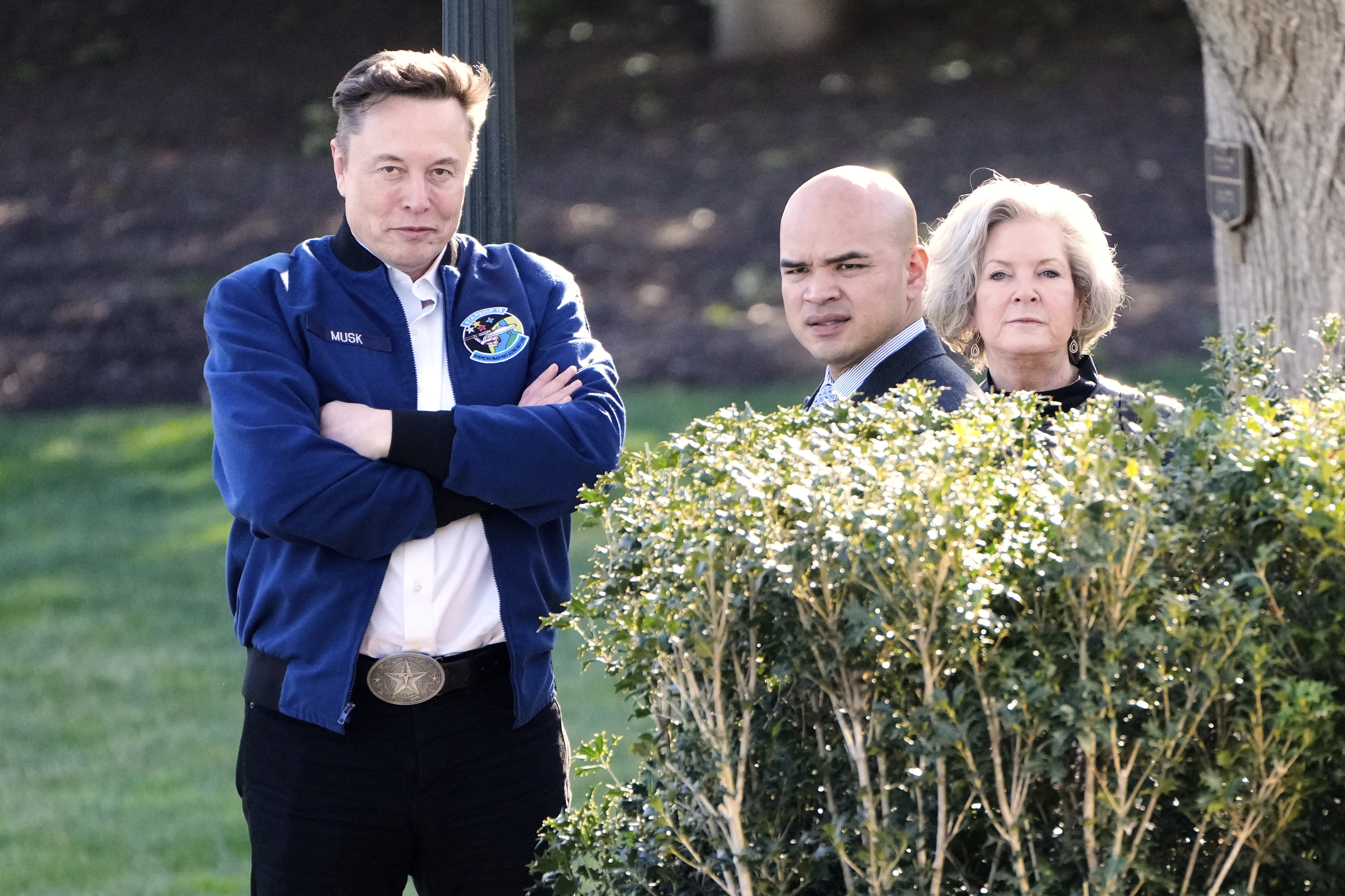 Elon Musk crosses his arms, next to Walt Nauta and Susie Wiles, standing on the White House lawn.