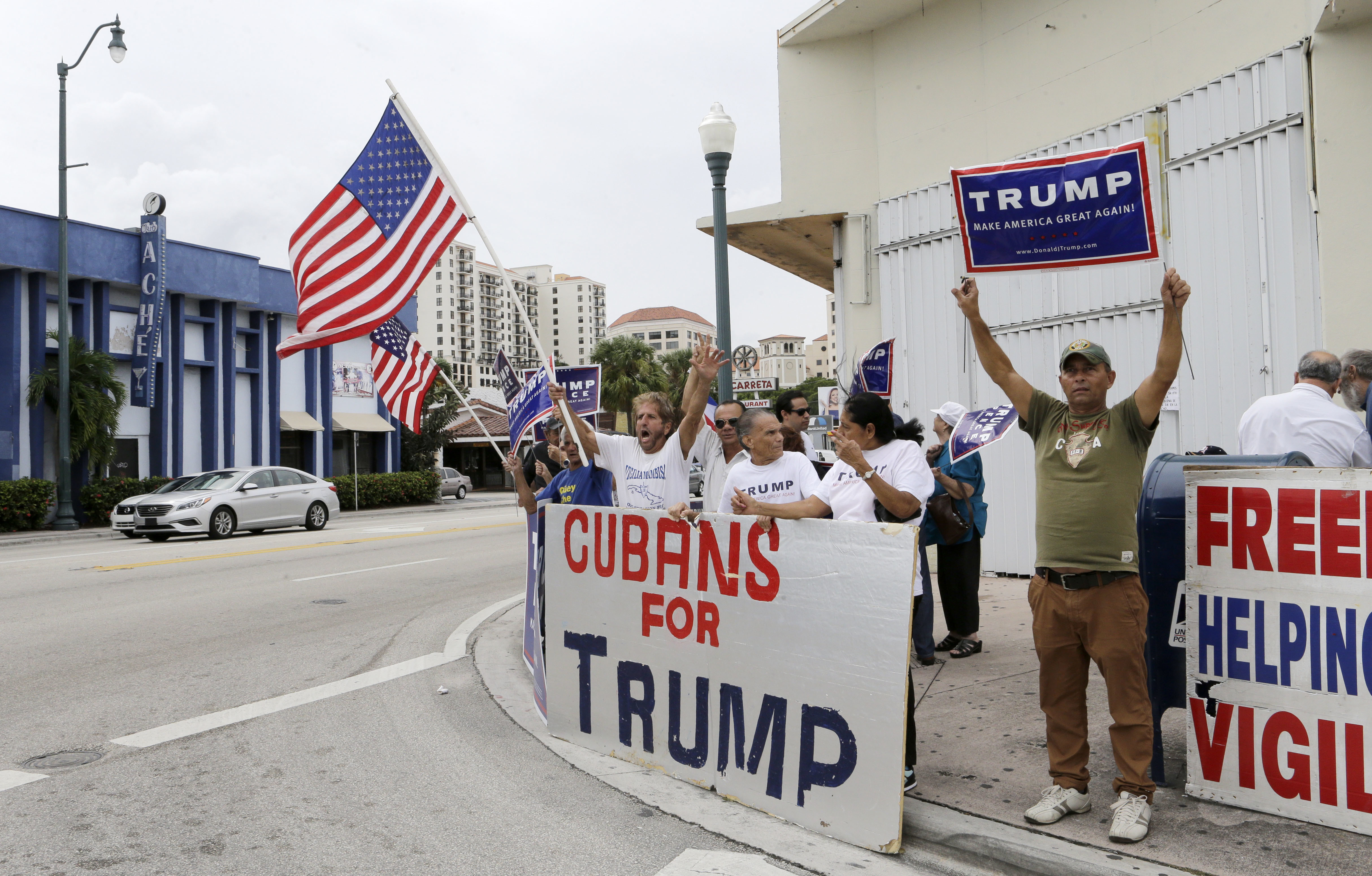 Protesters hold up banners on the side of the road reading, "Cubans for Trump" and wave US flags.
