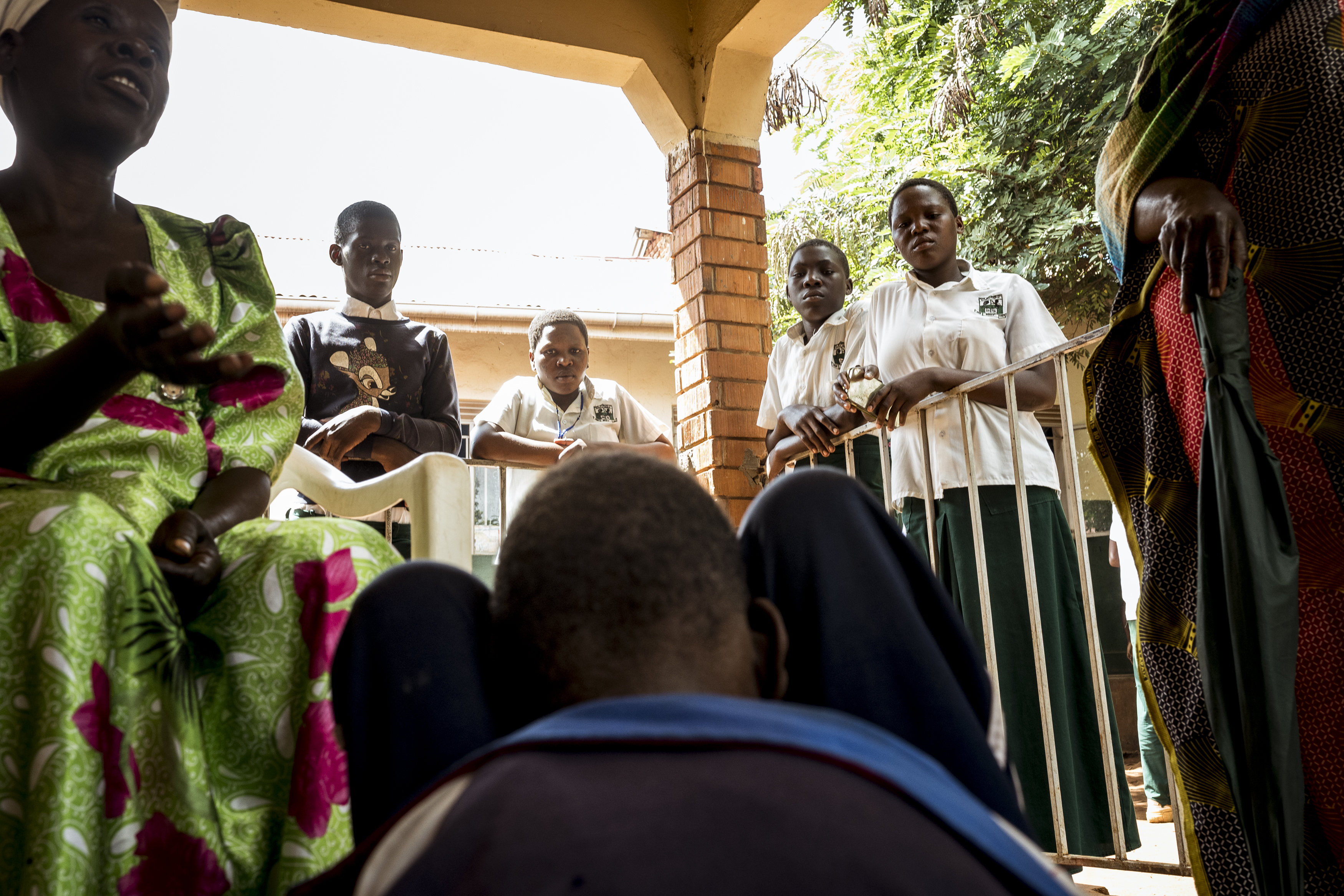 Buwenge, Uganda. 16-year-old Moses is ogled by local students as his mother, Evelyn Mbwali explains his story to a health worker at a local outreach clinic. Moses was brought to the outreach by his mother who was worried about his lack of mobility. Severely malnourished, and epileptic, because of poor positioning he has developed a curved spine and contractures in his knee joints. Community awareness and acceptance surrounding disability, whilst increasing, is still at a level whereupon there is a great deal of stigma attached. Families are often ostracised from their communities; parents will abandon children born with disability and in some cases 'mercy killings' are performed. Local community-based organisations and NGO's work tirelessly on educational programs that look to dispel these myths, incorporating information sessions into outreach programs in the hope the knowledge will be taken back to local villages and the information shared. [Christopher Hopkins/Al Jazeera]